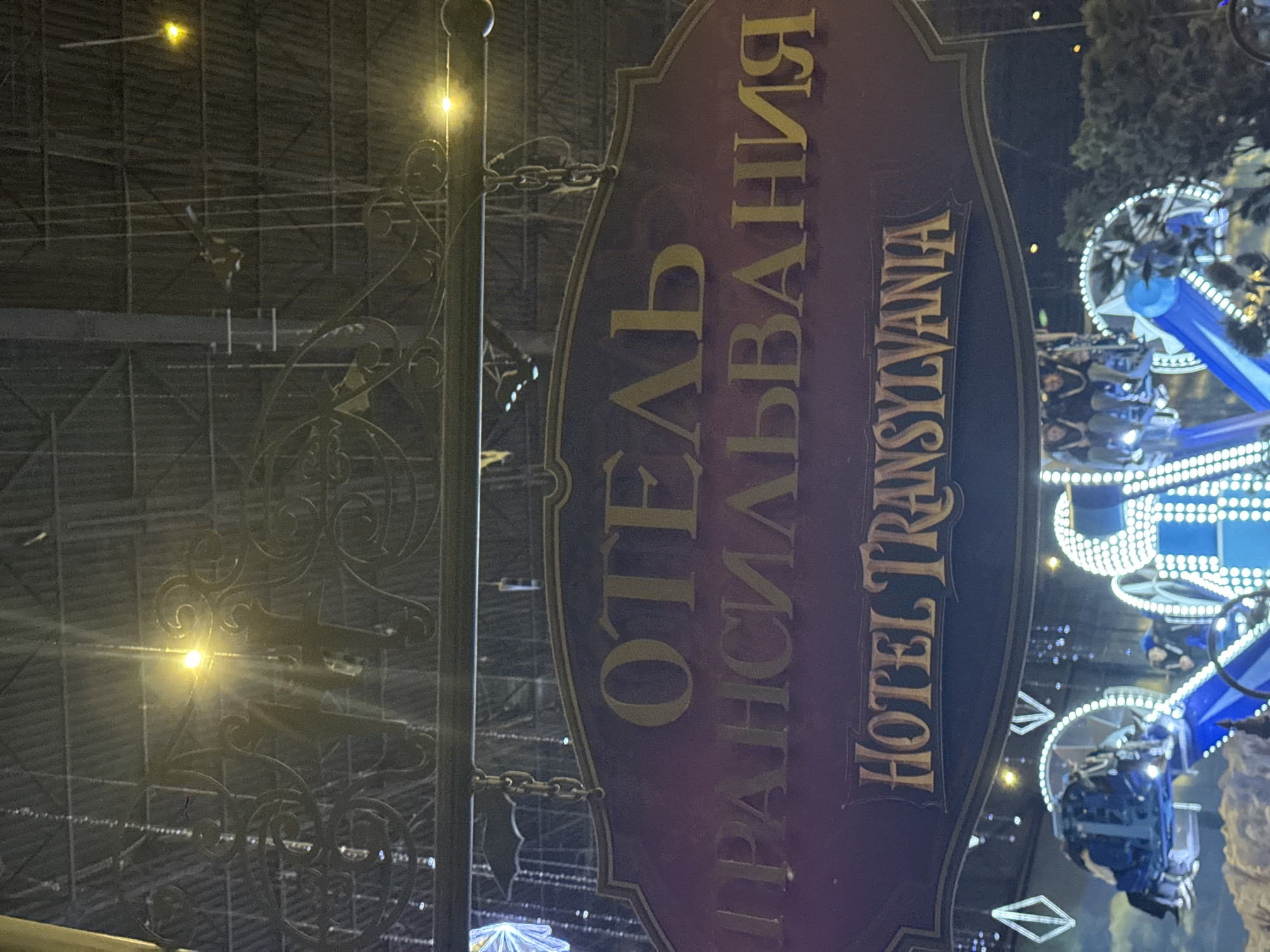 The image depicts a sign for "Hotel Transylvania" in both Russian and English. The sign is mounted on a decorative metal post with scrollwork. In the background, there is an amusement park ride with bright white lights and blue accents. People can be seen on the ride. The location is the Island of Dreams theme park in Moscow, Russia.