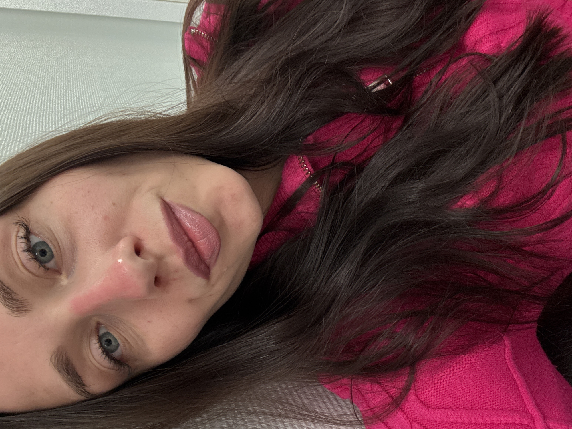 A close-up, rotated portrait shows a young woman with long, dark brown hair and fair skin. Her face, seen from an overhead perspective and tilted, occupies the left side of the frame. She has clear blue eyes with dark eyelashes and eyebrows. Her lips are made up with a pinkish-mauve lipstick, and she has a neutral, calm expression with a slight, subtle smile. The bridge and tip of her nose appear notably reddened.
Her dark, wavy hair is spread around her head and over her shoulder, contrasting with the vibrant fuchsia or hot pink knitted garment she is wearing. This garment has a textured pattern and features silver zipper teeth visible among her hair. She appears to be lying down, with her head resting on a light, textured surface that could be a pillow or a light-colored headboard/wall. The background behind her is a light, off-white or pale gray wall with a subtle vertical striped texture. The lighting is soft and diffused, suggesting an indoor setting. There is no visible text or specific environmental cues to confirm the Saint Petersburg, Russia context.