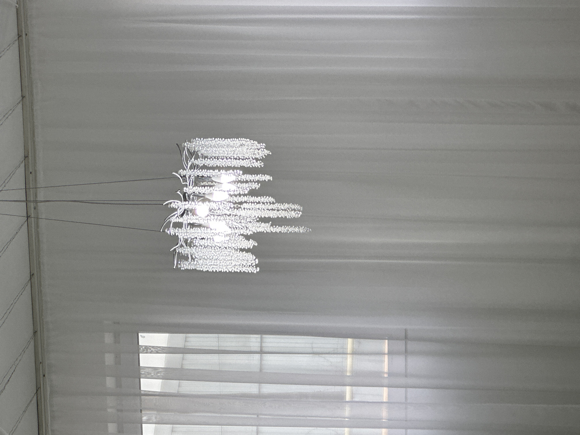 The image captures a chandelier hanging from a ceiling, possibly within a tent or event space. The ceiling is draped with fabric, creating a textured appearance. Below the chandelier, a window with blinds is visible.