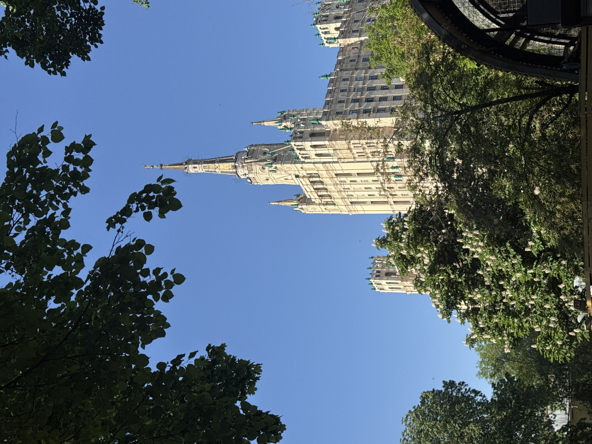 The image captures a low-angle view of one of the Seven Sisters skyscrapers in Moscow, Russia, framed by trees. The building is tall and features a distinct spire. The sky is clear and blue, with tree foliage visible in the foreground. A portion of a dark, possibly metal, structure is present at the right side of the frame. The overall scene is taken during daylight.
