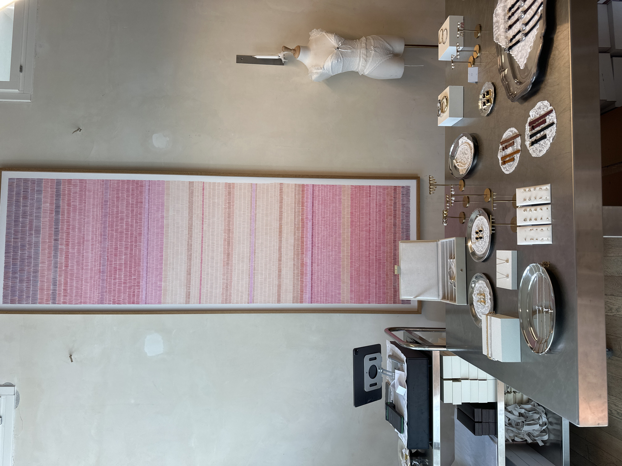 The image depicts the interior of a retail space, possibly a jewelry or clothing boutique. The setting is likely Tel Aviv, Israel, although this is not explicitly stated.
Key elements include:
- A large, framed artwork with horizontal stripes in shades of pink and purple.
- A headless, white mannequin torso mounted on the wall.
- A display table with various jewelry items arranged on trays and stands. The table also holds several product boxes.
- A tablet is mounted next to the display table.
The environment is minimalistic with light-colored walls. The focus of the image is on the presentation of the merchandise and artwork within the retail space. There are no people visible in the frame.