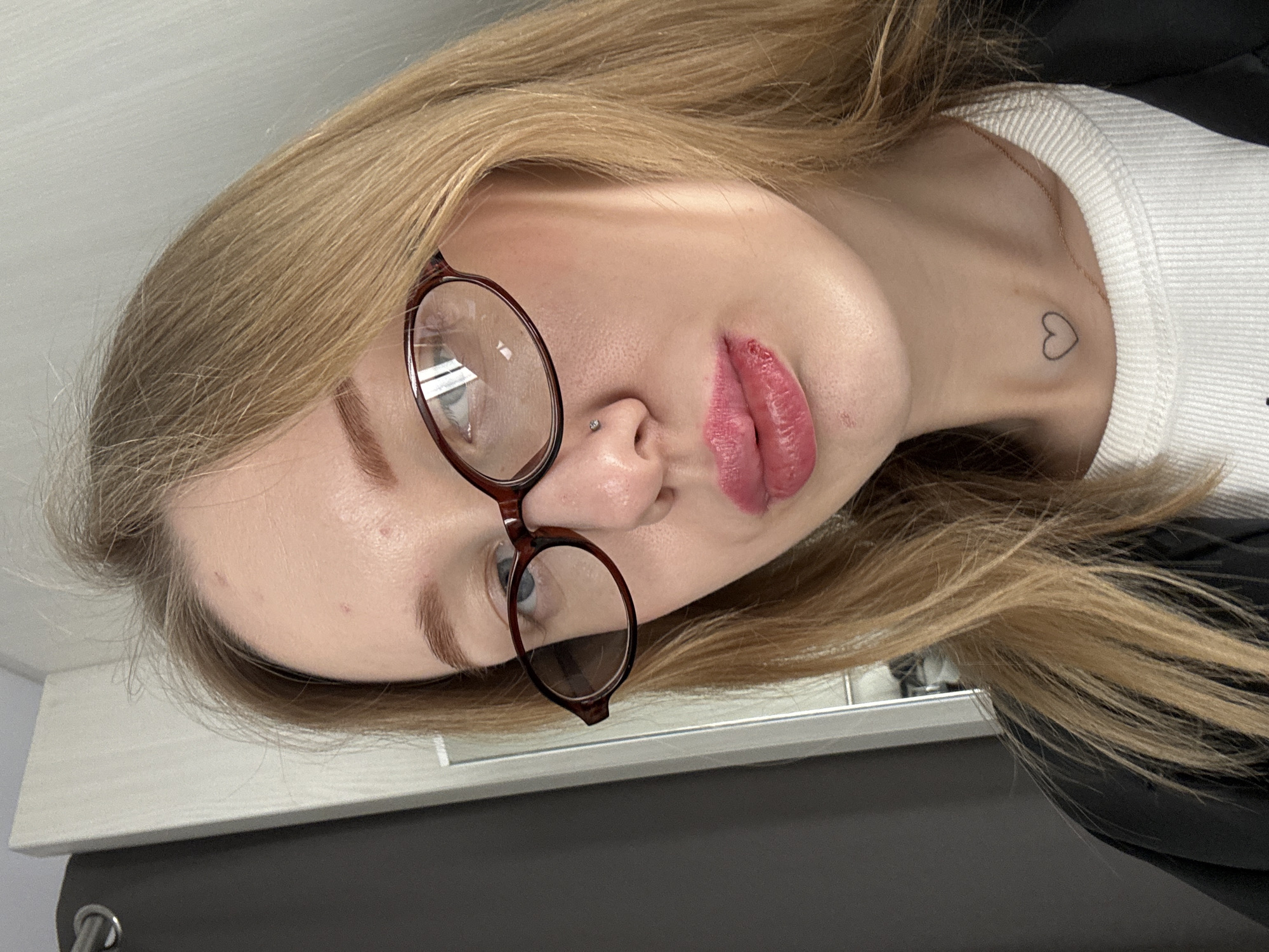 A female subject is depicted in a close-up, upward-angled self-portrait, captured indoors in Rostov-on-Don, Russia. The subject has long, light blonde hair, parted on the right. She wears dark reddish-brown, oval-framed eyeglasses, through which her blue eyes are visible, directed slightly upwards and to the left. A small, clear stud is present in her left nostril. Her lips are full, enhanced with pink lipstick, and exhibit some texture on the lower lip. A small, outlined heart tattoo is visible on her lower neck, above a white ribbed crew-neck top. She is also wearing a dark-colored outer garment, possibly a jacket or shirt, over the white top. The immediate background on the left consists of a dark gray curtain or fabric with a silver curtain ring. To the right, a light-colored wall or furniture panel is visible, separated from the curtain by a narrow vertical reflective strip, potentially a mirror or glossy furniture element.