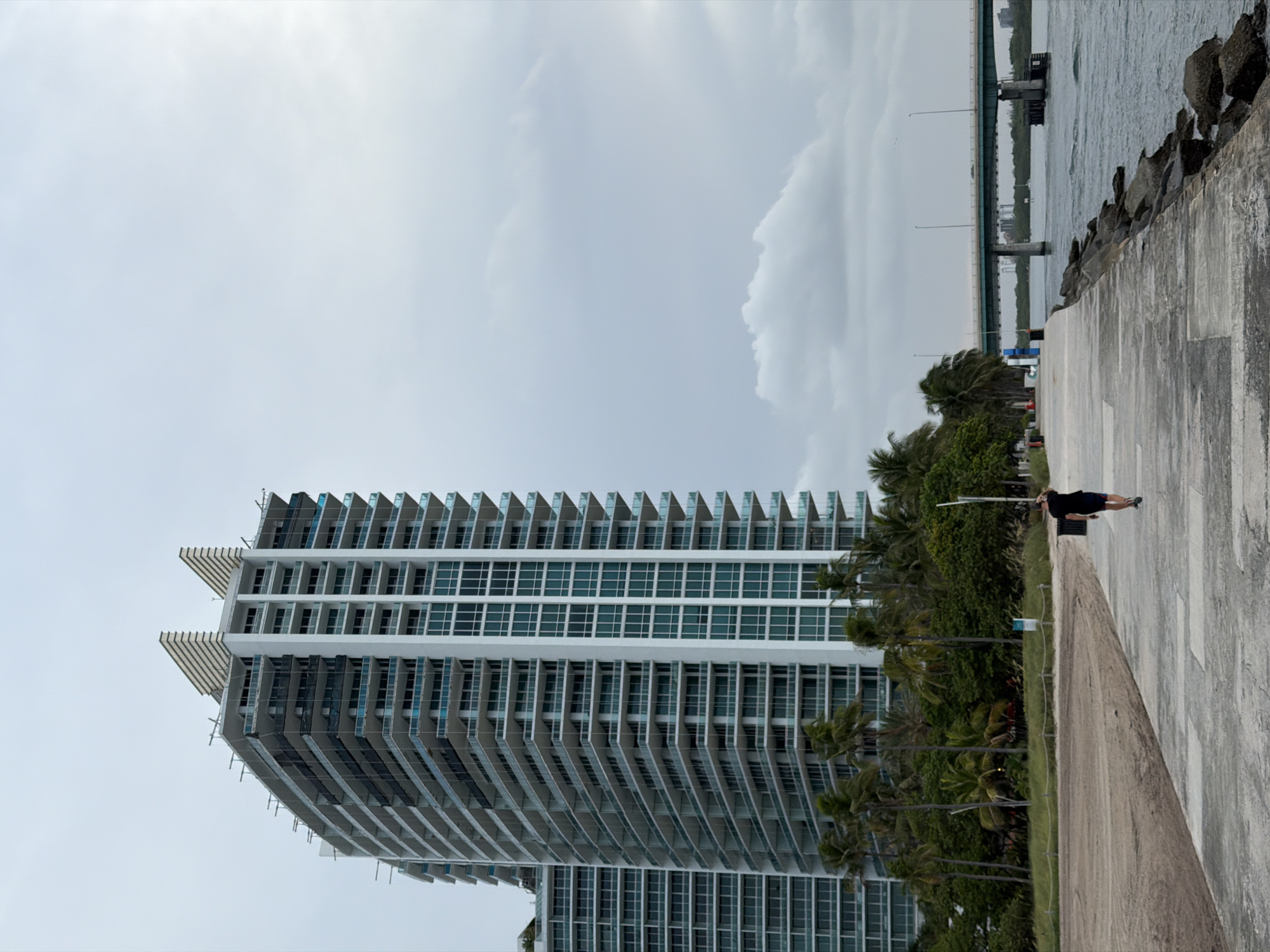 A full shot captures a person walking on a concrete pier, facing away from the camera, with a large, modern building in the background. The building, featuring numerous balconies and glass windows, is tall and appears to be under construction in some sections. Palm trees are scattered around the base of the building, and a beach is visible to the left. The pier extends towards a body of water on the right, where a bridge spans across it in the distance. The sky is overcast, suggesting a cloudy day. The scene is set in Bal Harbour Village, United States of America.