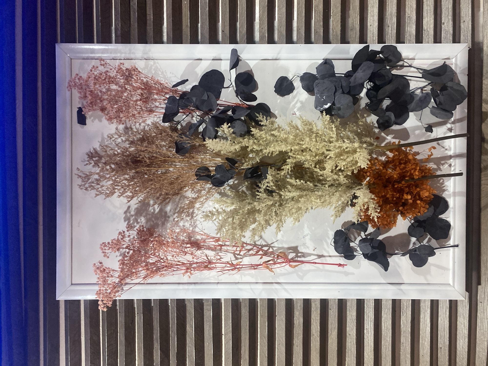 A framed arrangement of dried flowers is mounted on a wall with horizontal wooden slats. The frame is white and rectangular. Inside the frame, there are several bunches of dried flowers. On the left side, there are two bunches of pink, feathery flowers. In the center, there is a large bunch of pale beige, bushy dried grass. Behind this, and slightly to the right, is a bunch of dried, spiky brown foliage. In the lower center, there is a small bunch of vibrant orange dried flowers. To the right of the beige grass and extending downwards, there are several sprigs of dark, almost black, foliage with rounded leaves. The background of the frame is white. The wall behind the frame consists of parallel wooden slats, with a dark blue band at the very top. The scene appears to be indoors, possibly in a residential or commercial setting in Ivanteyevka, Russia, given the reference to the city. No people or animals are present.