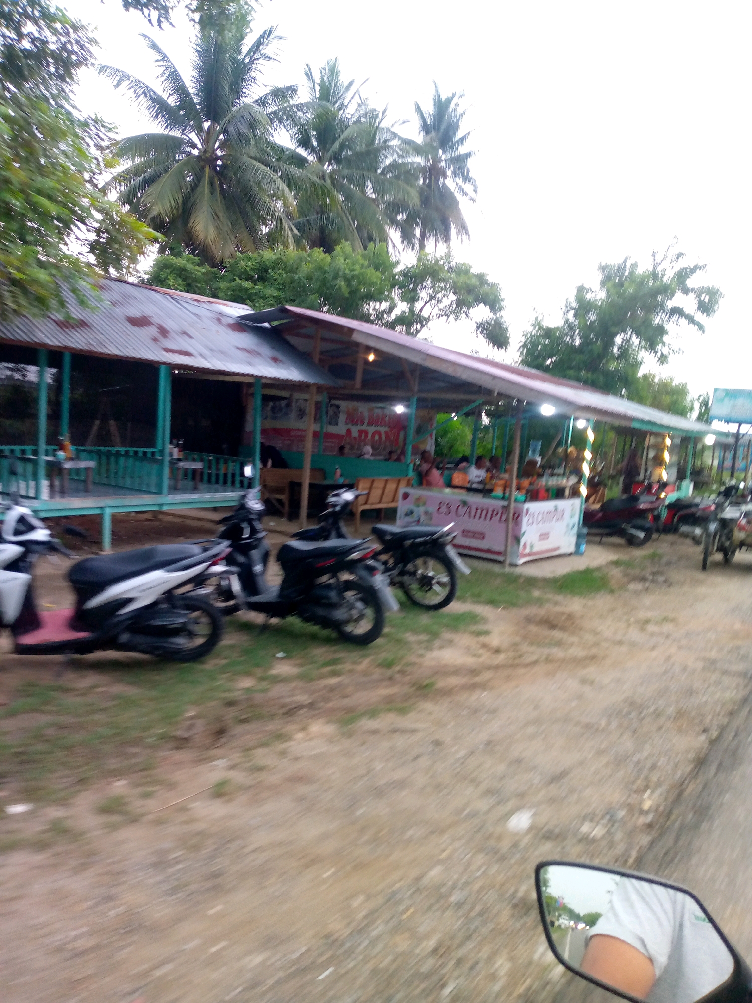 This is a slightly blurred, eye-level, medium shot of a street scene in Keudearon, Indonesia. On the left, there is a food stall with a corrugated metal roof supported by wooden posts. The stall is open to the front and has a green railing. Several tables and chairs are visible inside. In front of the stall, parked on the ground, are several scooters. One white scooter is in the foreground, and behind it, a black scooter is parked. In the background, there are palm trees and other vegetation. The ground appears to be a mixture of dirt and grass. On the right side of the image, a motorcycle's rearview mirror reflects a person wearing a light-colored t-shirt and the road ahead. The road is paved and has other vehicles visible in the reflection. The overall impression is that of a casual outdoor eating area alongside a road.