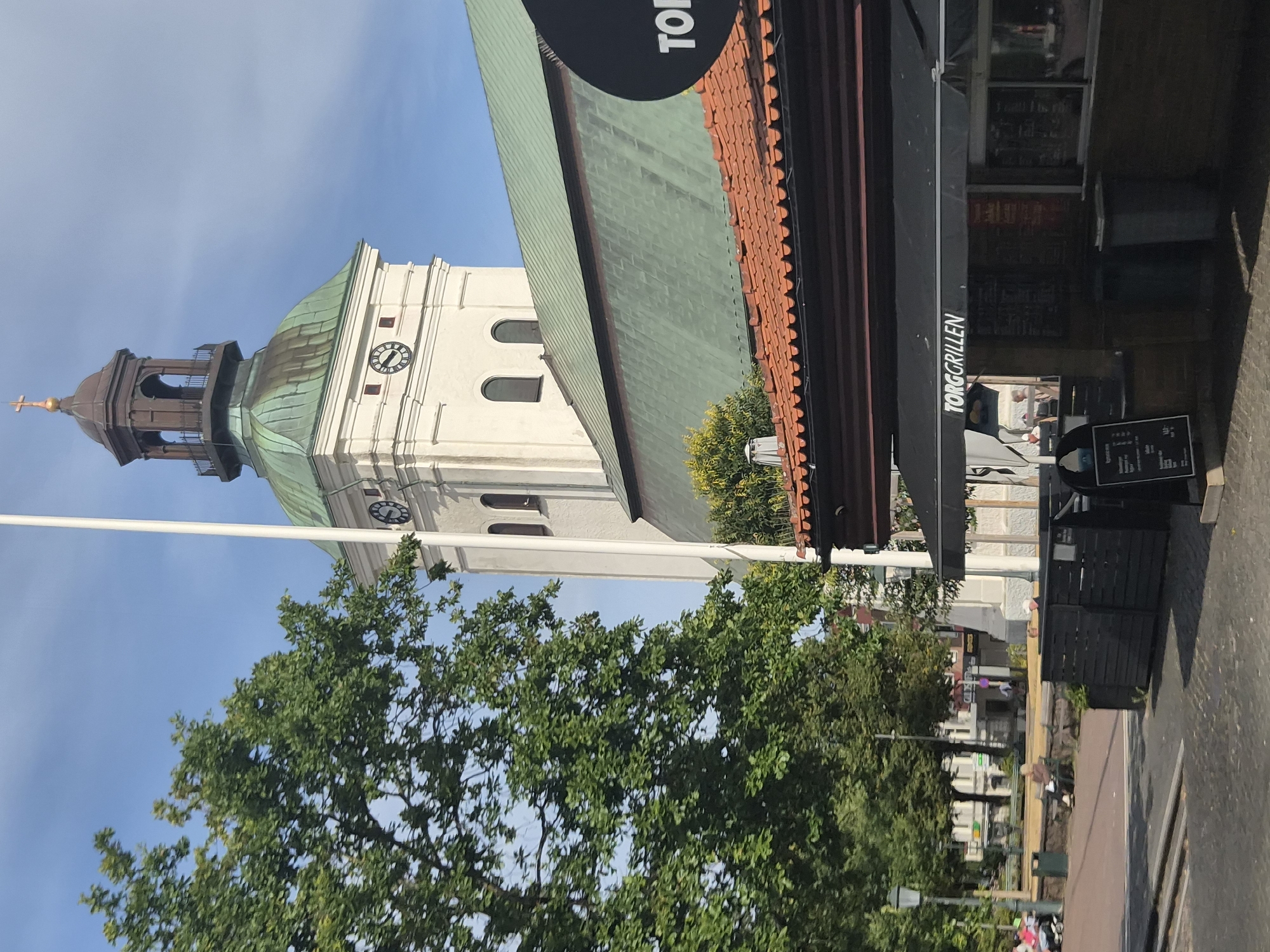 The image depicts a street scene in Varberg, Sweden, with a prominent white church tower featuring a green copper dome and a dark bell tower topped with a golden cross. A large, mature green tree is positioned to the left of the church. In the foreground, a covered outdoor dining area for "TORGRILLEN" is visible, with a black awning displaying the restaurant's name in white lettering. Several tables and chairs are arranged under the awning, and a menu board is placed on an easel. People are seated at tables and walking in the background. The sky is clear and blue.