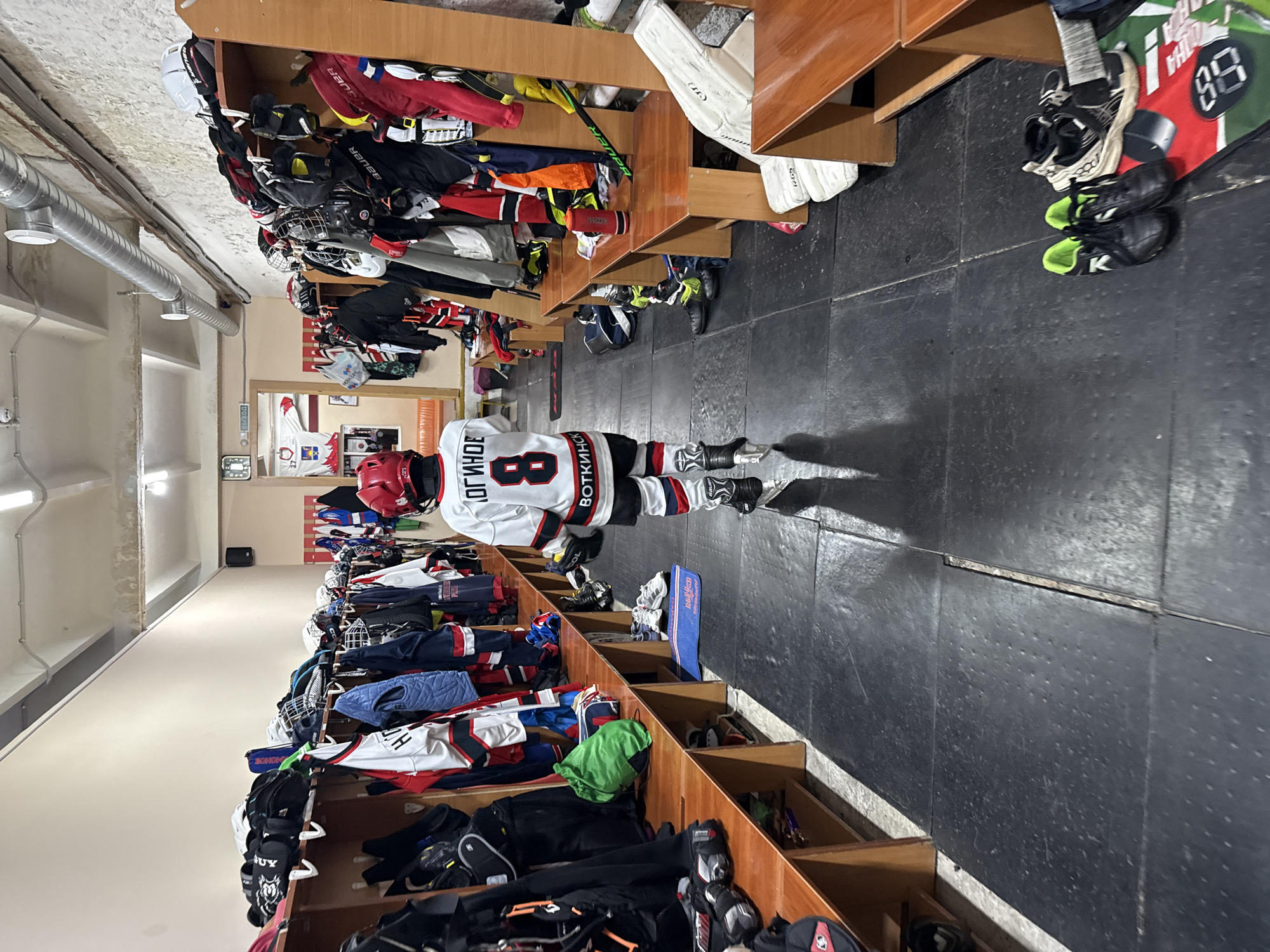 The image depicts the interior of a locker room, likely at a sports facility in Votkinsk, Russia. In the center, a young person wearing a white hockey jersey with the number "8" and Russian lettering, along with full hockey gear including skates, a helmet, and gloves, is walking away from the camera. The locker room is equipped with wooden benches and numerous shelves filled with hockey equipment, including helmets, pads, bags, and clothing. Several pairs of skates and other footwear are visible on the floor and on the shelves. The floor is covered with black rubber matting. The environment appears functional and utilitarian, suggesting a space used for athletic activities. The overall impression is one of readiness or a post-activity scene within a sports context.