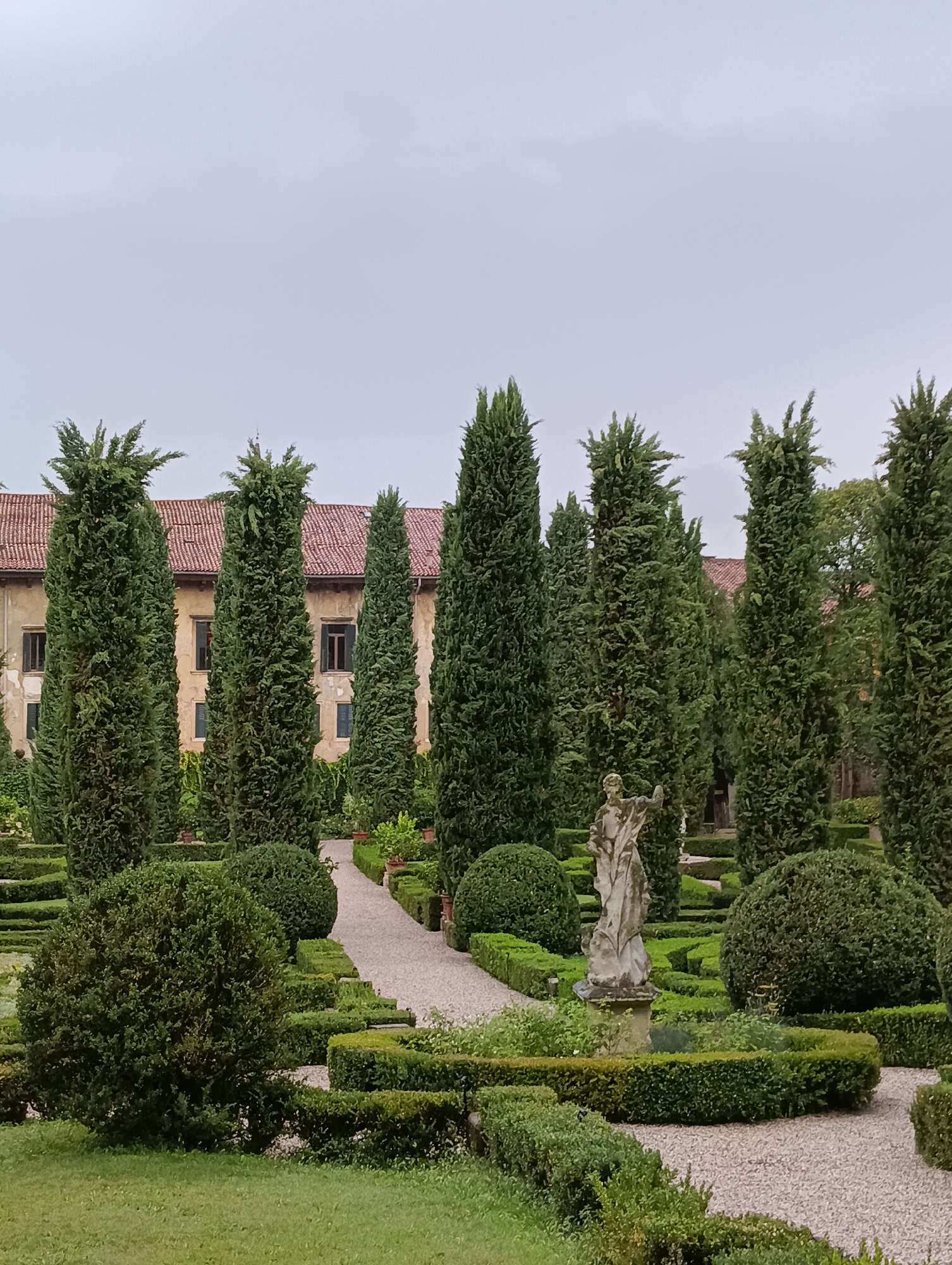 The image depicts a formal garden with a stone statue of a figure in the foreground, likely in a classical or baroque style. The statue stands on a pedestal within a meticulously manicured garden featuring neatly trimmed hedges forming geometric shapes and pathways. Tall cypress trees are interspersed throughout the garden, their dark green foliage contrasting with the lighter green of the hedges and grass. In the background, a building with a red tiled roof and light-colored walls, featuring windows and shutters, is visible. This style of architecture and garden design is characteristic of historical estates and villas, often found in regions like Veneto, Italy. The location is likely a historical villa garden in Verona, Italy, given the architectural style and landscaping typical of the area. The sky is overcast, suggesting a cloudy day. The overall scene evokes a sense of historical grandeur and horticultural artistry.