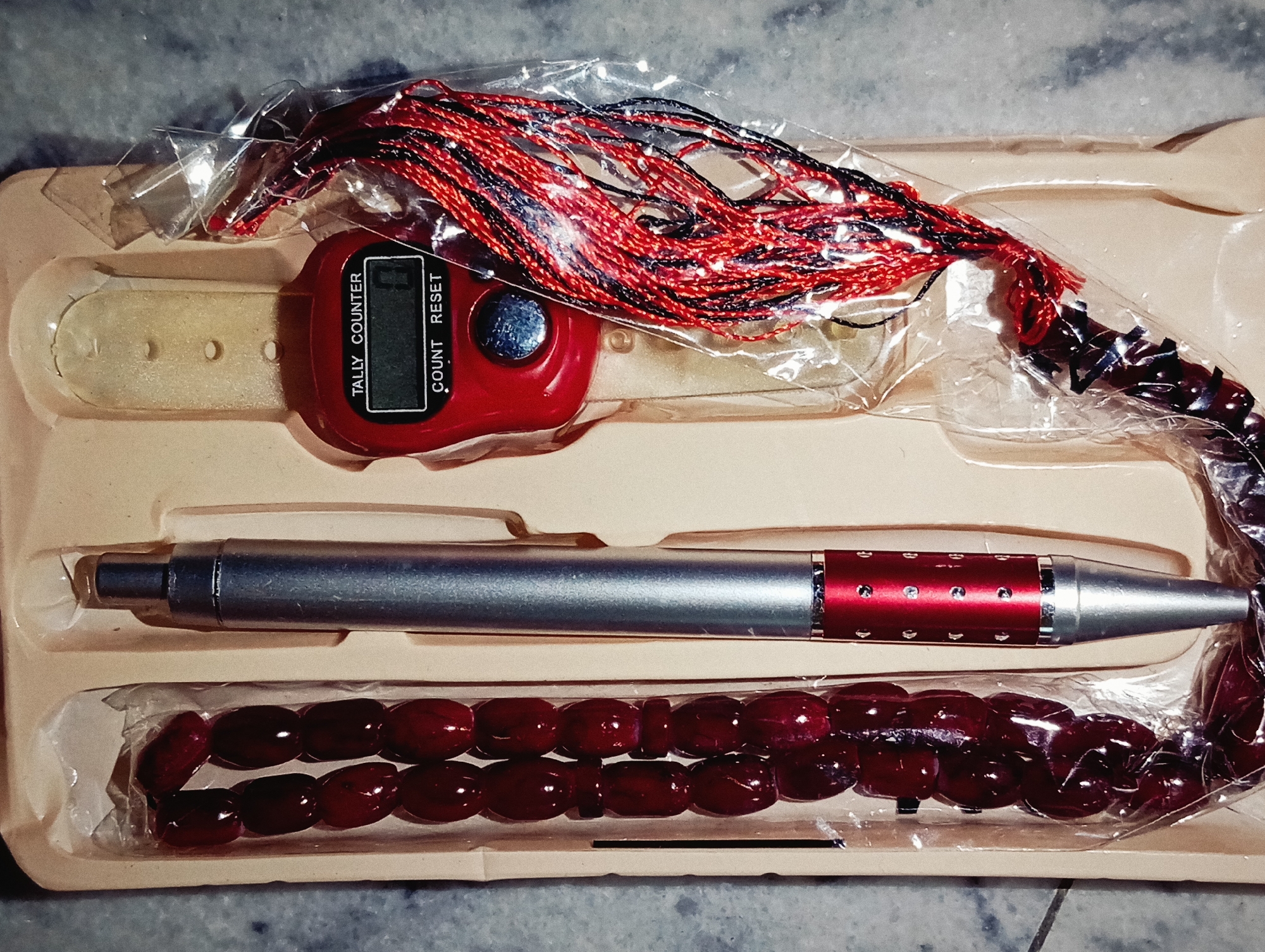 An overhead shot shows a red digital tally counter with a wristband, a metallic pen with red accents, and a strand of maroon prayer beads, all arranged in a light brown plastic tray. The tally counter's display shows "0". The pen has a silver body and a red section with small circular designs. The prayer beads are oval and have a glossy finish. A string of red, black, and metallic threads is attached to the tally counter. The background is a gray, speckled surface. The objects are presented in a clean, organized manner within the tray.