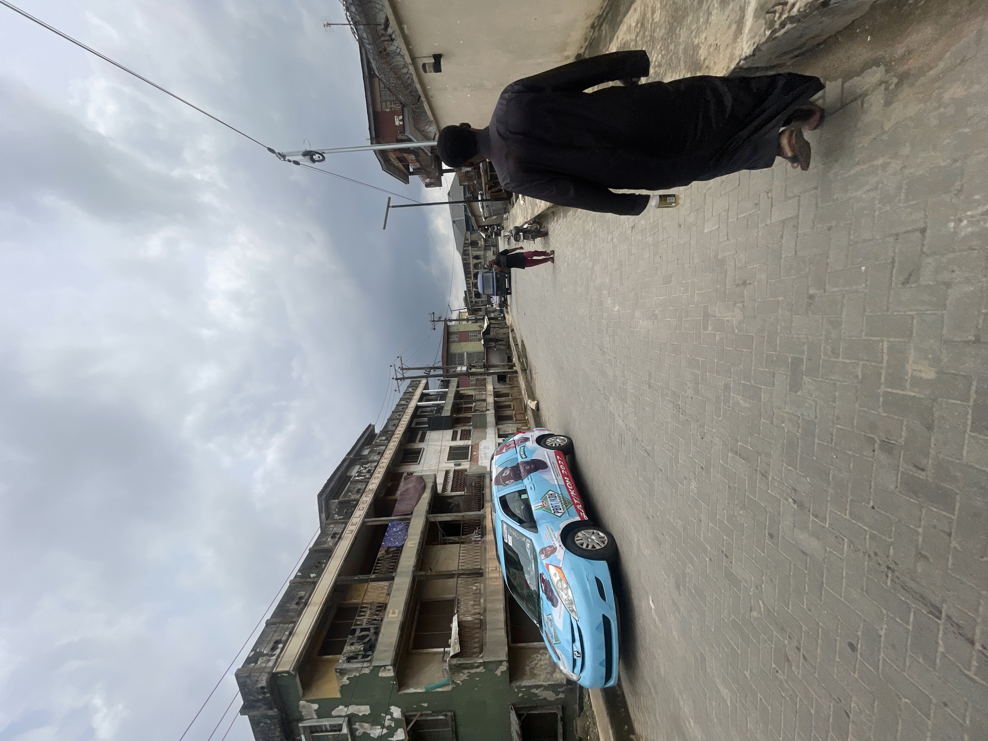 A man in a long black robe walks away from the camera down a narrow, paved street in Akowonjo, Nigeria. He is carrying a bottle in his right hand and wearing brown sandals. To his left, a light blue minivan is parked on the side of the road. The minivan is covered in colorful political advertisements. In the background, there are several multi-story buildings with balconies and laundry hanging out to dry. The sky is overcast with heavy clouds. The street appears to be a residential or commercial area with a mix of old and new construction.