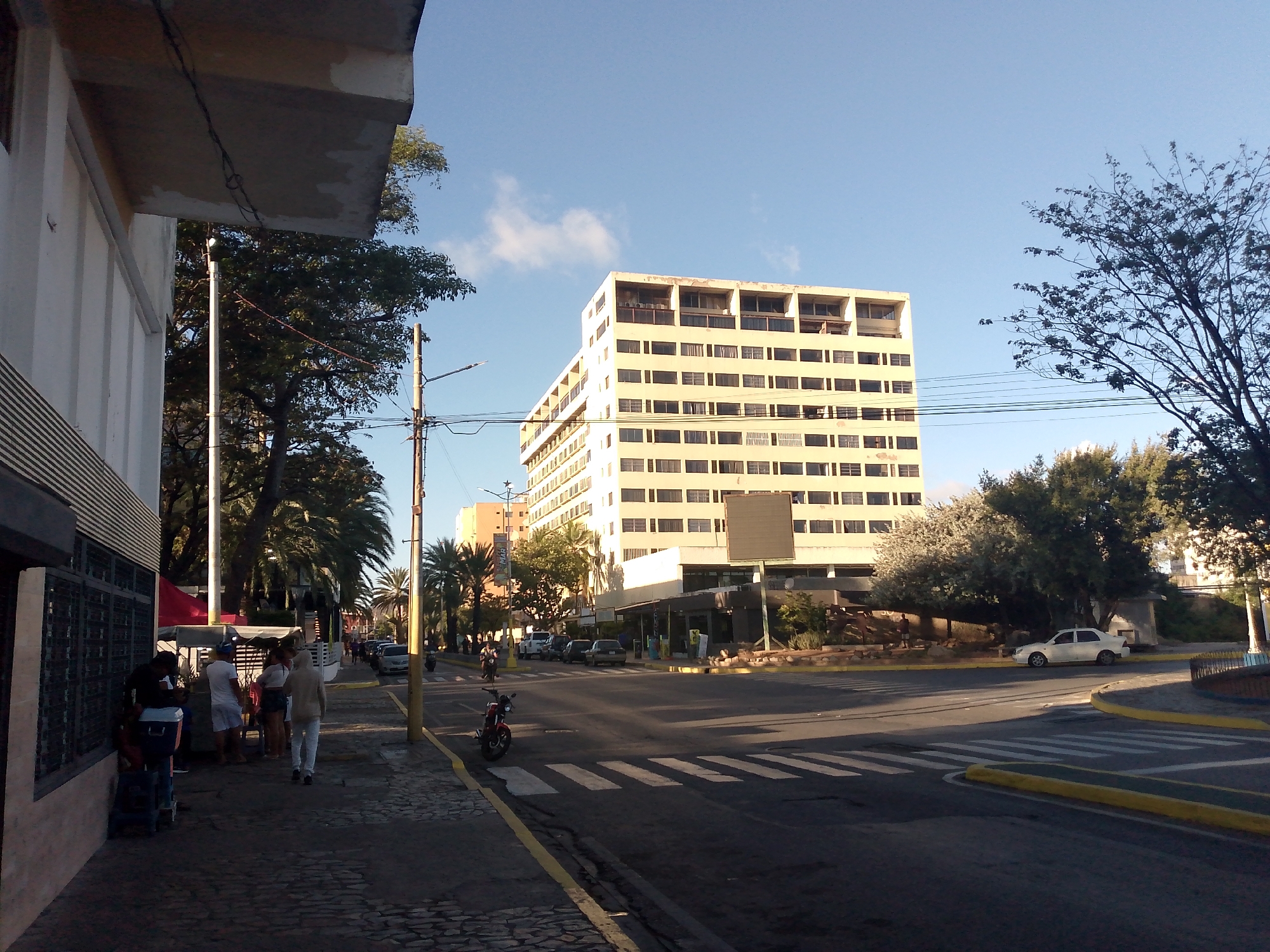The image depicts a street scene in Juan Griego, Venezuela, on a sunny day. The main subject is a large, light yellow or beige building with multiple floors and evenly spaced windows. The building appears to be near a street corner.
In the foreground, on the left side of the frame, people are gathered near a building with horizontal stripes along its lower portion. One person is walking away from the group, heading towards the crosswalk. On the street, there are several vehicles, including cars and motorcycles. A red motorcycle is parked on the side of the road near a yellow curb.
The street has zebra-striped crosswalks. The environment includes trees and vegetation, suggesting an urban setting with greenery. The sky is blue with a few scattered clouds. Utility poles and wires are visible above the street.