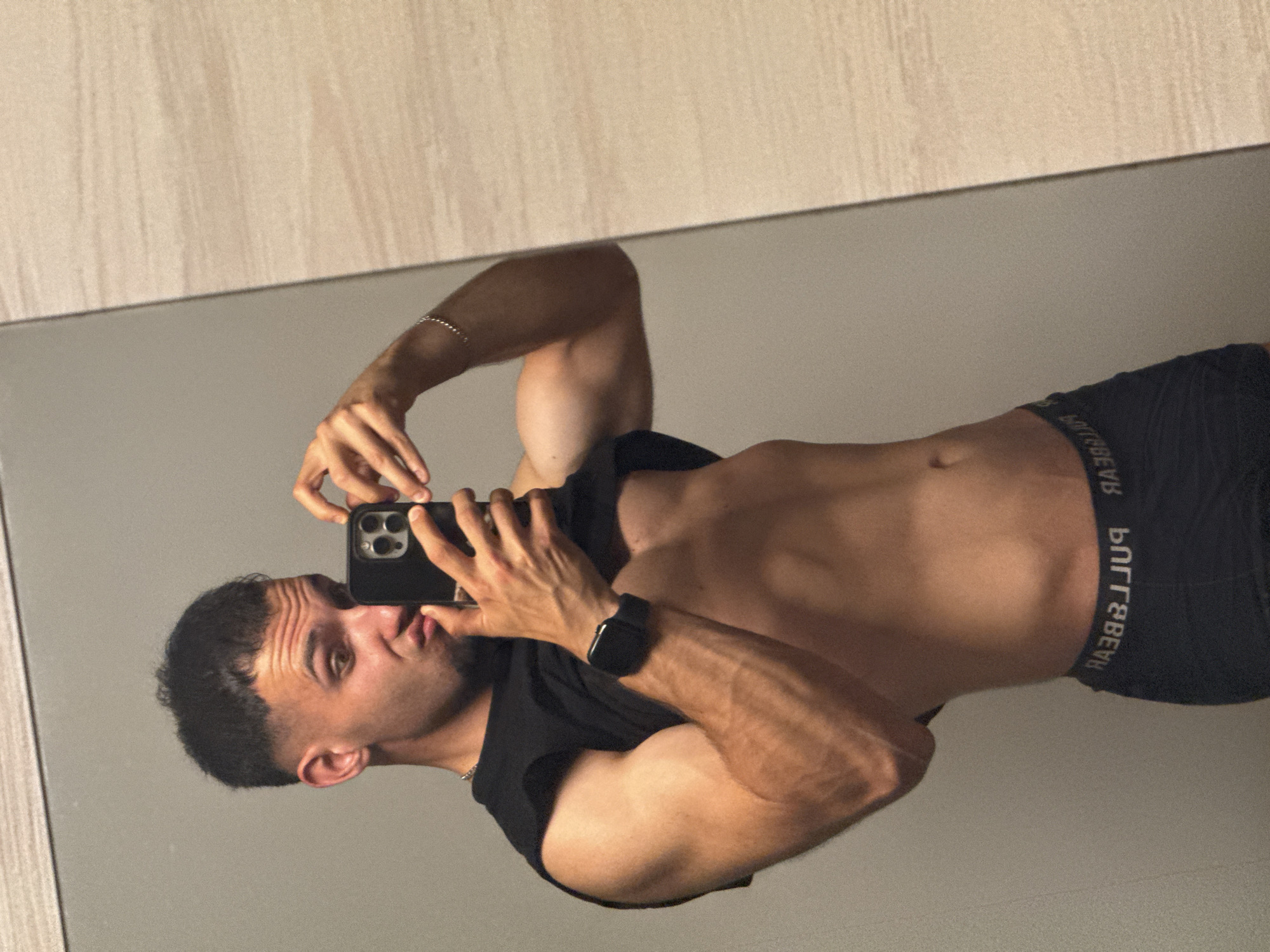 The image displays a man taking a mirror selfie indoors. He is reflected from the waist up, shirtless, with a black t-shirt pulled up to his chest. He wears black boxer briefs, a black smartwatch on his left wrist, and a thin silver bracelet on his right wrist. He holds a smartphone with multiple camera lenses in both hands, angled towards himself, capturing his reflection. His head is turned slightly to the left, and he has a casual expression with slightly puckered lips. He has short, dark hair and light stubble. The man appears to be physically fit.
The background visible in the mirror consists of a light-colored, possibly wooden, textured surface at the top, likely a ceiling or an upper part of a wall, and a plain light gray wall or surface below it. The lighting is soft and even, suggesting an indoor setting without harsh direct light.
Visible text includes the brand "PULL&BEAR" mirrored on the waistband of his boxer briefs. No specific location cues are discernible from the image itself to confirm the context of Shumen, Bulgaria.