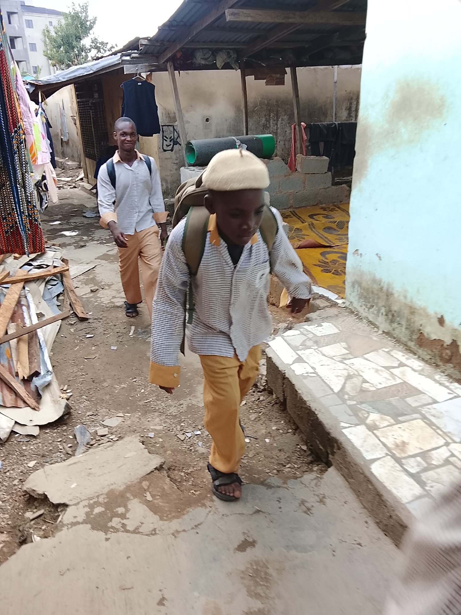 Two young males are walking on a dilapidated city street in Abuja, Nigeria. The foreground figure has a backpack and a cream-colored hat, wearing a long-sleeved collared striped shirt with orange cuffs and orange pants, and sandals. The figure behind has the same outfit except for the hat. They are walking on a broken stone paved street. To the right is a cement block wall and behind that is a corrugated steel roof. To the left is a structure with hanging clothes and other items. The street is littered with trash and debris. A multistory apartment building can be seen in the upper left.