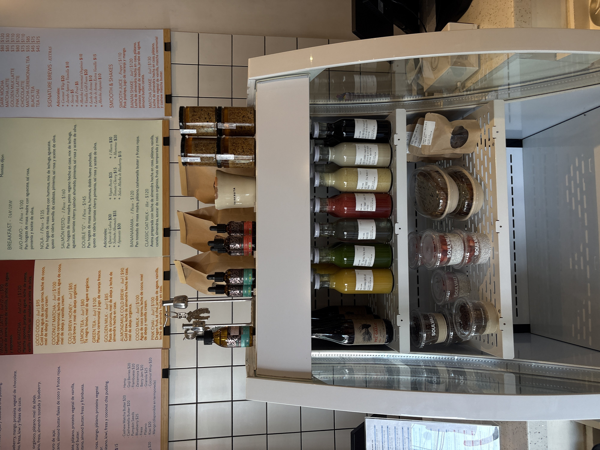 The image displays a well-organized interior of what appears to be a health food cafe or store. The main subjects are a white display refrigerator filled with various products and a detailed menu board mounted next to it. The background features a white tiled wall.
The display fridge contains multiple shelves stocked with bottled beverages, including juices, milks, and kombuchas, some prominently labeled "SOULVALI" and "PEZ GALLO". Several paper bags and clear plastic containers, also branded "SOULVALI", holding items like chia puddings or prepared meals, are visible. Another brand, "THISFRUTA", is also seen on some packaging.
To the left of the fridge, a comprehensive menu board lists a wide array of food and drink options in Spanish. Drink categories include "DARK MOCHA", "MATCHA LATTE", "GOLDEN MILK LATTE", "PINK CHAI LATTE", "CHOCOLATTE", "MATCHA CEREMONIAL TEA", "BLACK TEA", and "TEA CHAI," each with two price points (e.g., $80, $120). The "SIGNATURE BREWS - EXTRAS" section details iced beverages like "VANILLA OAT LATTE" ($90), "OLIVE MATCHA" ($120), and "GREEN TEA" ($100), along with various "Adicionales:" (add-ons) such as "Coconut Whip" ($20), "Stevia" ($5), and "Matcha Japonés" ($10). The "SMOOTH & SHAKES" section lists "SMOOTH JUICE - Frappé" ($110), "BANAN SHAKE" ($120), and "MATCHA SHAKE" ($130), with descriptions of their ingredients.
The "BREAKFAST - 7AM-12PM" menu features savory toasts like "AVO ARVO" ($100), "NOLA" ($135), "SALMÓN PESTO" ($160), and "DOUBLE 'G'" ($145), along with sweet options such as "BANANARAMA" ($120) and "CLASSIC OAT MILK" ($120). Additional toppings like "Queso de Cabra" ($30) and "1 Huevo" ($30) are also listed. A separate section itemizes prices for various smoothie bowl ingredients and add-ons like "Cashew Matcha Butter" ($20), "Goji Berries" ($20), and "Granola" ($25).
The environment appears clean and well-lit, suggesting active business hours. No people are visible. The currency indicated is pesos ($), and the language is Spanish, consistent with the location context of Puebla, Mexico.