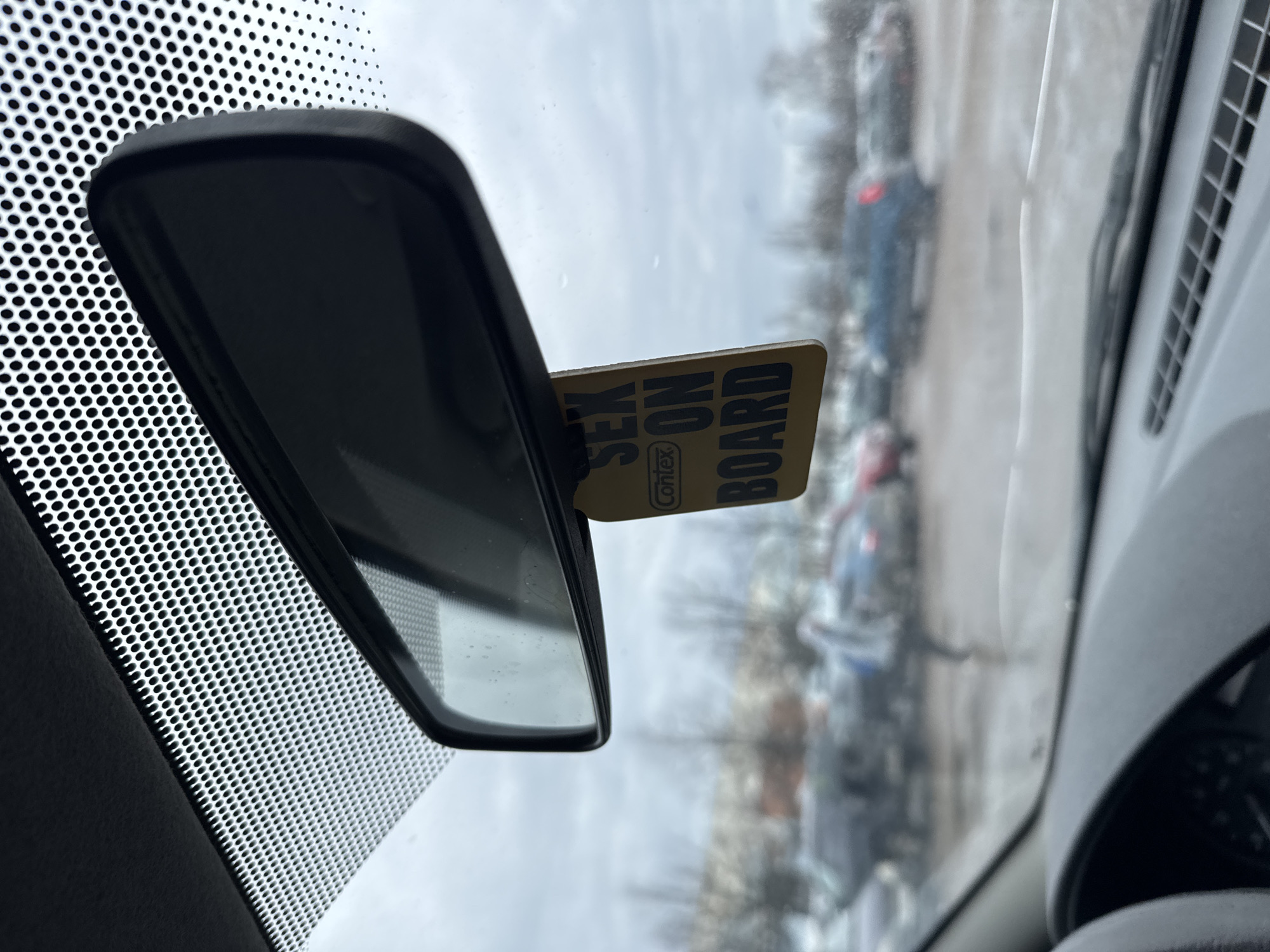 The image provides an interior view from a car in Chaikovsky, Russia, looking out through the front windshield. The car's rear-view mirror is prominently featured in the center-left, with its dark, reflective surface showing a blurred reflection of the car's interior and part of the windshield.
Attached near the top right edge of the rear-view mirror is a small, rectangular card. The card is light brown or yellowish with bold black text. Clearly visible on the card are the words "SEX ON BOARD", with the brand name "Contex" in a smaller, oval-shaped logo below "SEX".
The upper portion of the windshield features a distinctive, uniform pattern of small black dots on a white background, which is likely a factory-applied sunshade or frit band. To the right, a section of the car's interior, possibly the dashboard or a pillar, is visible with a grey textured surface and a partial view of an air vent.
Through the windshield, a blurred exterior scene is visible, suggesting an overcast daytime. Other vehicles are faintly discernible in the background, indicative of a parking lot or street. The lighting is soft and diffused, characteristic of cloudy weather. There are no people visible, and no specific activity is occurring.