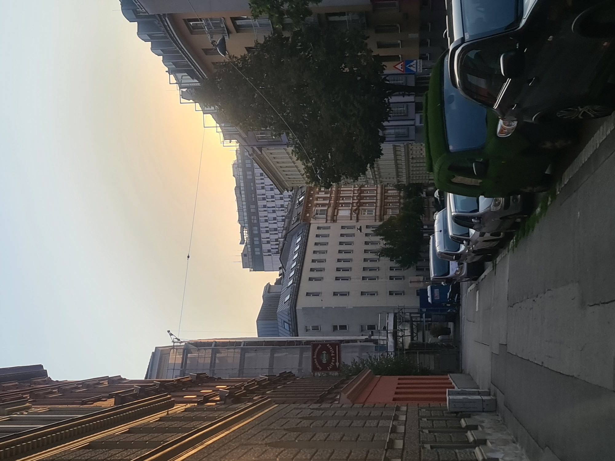 The image depicts a street scene in Vienna, Austria, at dusk. On the left, a multi-story building with ornate architectural details, possibly of historical significance, lines the sidewalk. The building is made of brick with stone accents and has arched window openings. To the right of this building, a smaller building with a red facade is visible, and further down the street, scaffolding surrounds a building under construction.
The street itself is paved with concrete slabs and is lined with parked cars on the right side. Several vehicles are visible, including a peculiar van covered entirely in green turf, resembling a bush. Other cars include a dark SUV, a light-colored hatchback, and several other vehicles further down the street.
In the background, a row of apartment buildings stretches along the street, with varying architectural styles and colors. Some buildings feature balconies and mansard roofs. The sky is a pale yellow and orange, indicative of a sunset, with the sun's glow diffused by the atmosphere. Trees are planted along the sidewalk in some areas, adding a touch of greenery to the urban landscape. A street sign with a pedestrian symbol and German text ("Schulweg") indicating a school route is also visible near the green van. The overall impression is a quiet, residential street in Vienna during the late afternoon or early evening.