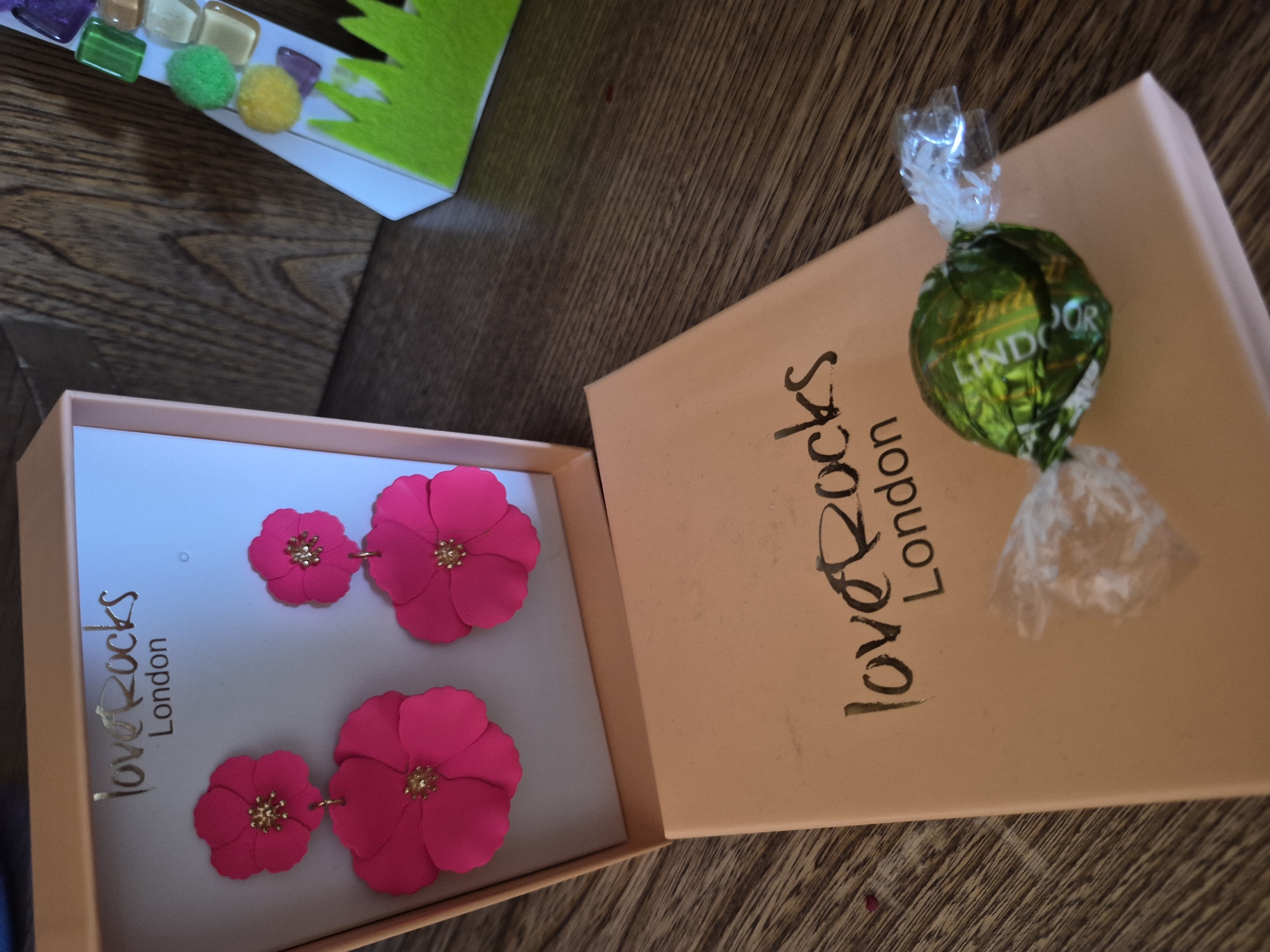 This image shows a close-up, overhead view of a gift box containing a pair of vibrant pink flower-shaped earrings. The earrings have small gold accents in the center of each flower. The box, also a light pink color, is open, revealing the jewelry inside. Outside the box, resting on the lid, is a green-wrapped Lindt chocolate truffle. The background is a wooden surface with a visible grain.
In the upper left corner of the image, a strip of colorful mosaic tiles and small pom-poms in green and yellow are visible, hinting at a crafting or decorative context. The brand name "love Rocks London" is printed in black script on both the inside of the box and on the outside lid. The overall impression is of a gift being presented, possibly for a special occasion, given the decorative items and the chocolate. The lighting appears to be indoor, and there are no strong indicators of the time of day or specific weather conditions. The setting is domestic or retail, given the objects and presentation.