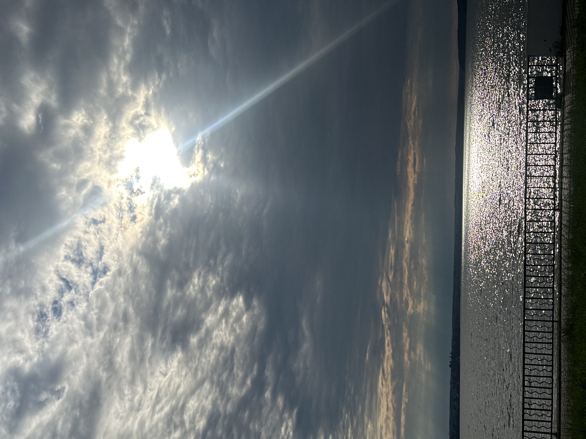 This image depicts a body of water, likely a lake or reservoir, with a sunlit path of shimmering reflections stretching across its surface. The sky above is dramatic, filled with a mix of dark, textured clouds and brighter patches where the sun is breaking through, casting a powerful sunbeam diagonally across the scene. In the foreground, a dark, ornate metal fence runs horizontally, separating a grassy area from the water. The horizon line shows a distant shoreline with trees and a few structures. The overall atmosphere is one of natural beauty with a touch of moody weather. The location is identified as Votkinsk, Russia. There are no people visible in the image.