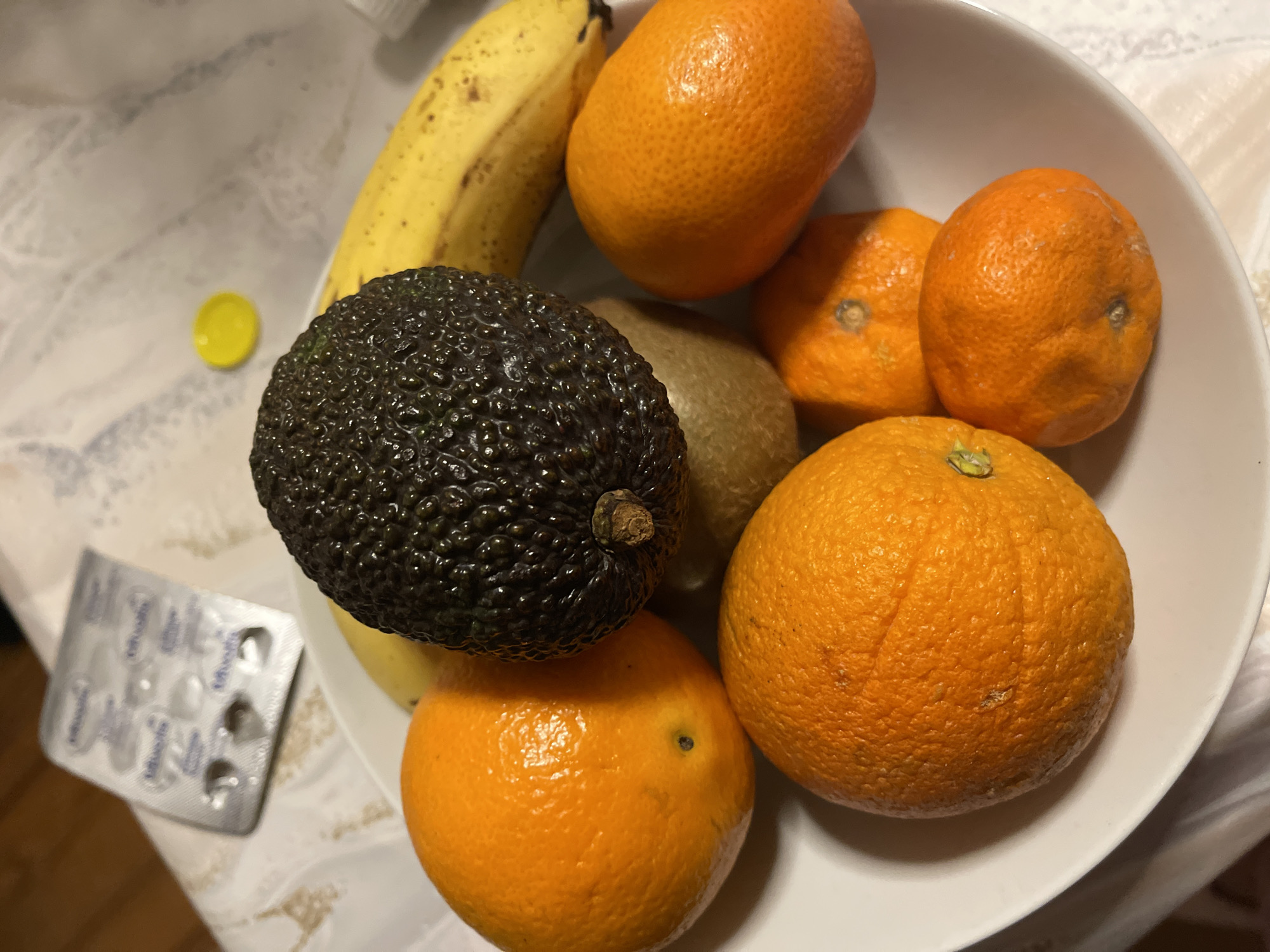 A top-down, close-up shot of a white bowl filled with various fruits, taken indoors. The fruits include a banana, an avocado, a kiwi, and several oranges or tangerines. A yellow bottle cap and a blister pack (possibly of medication) are visible on the side of the bowl. The location is not discernible from the image.