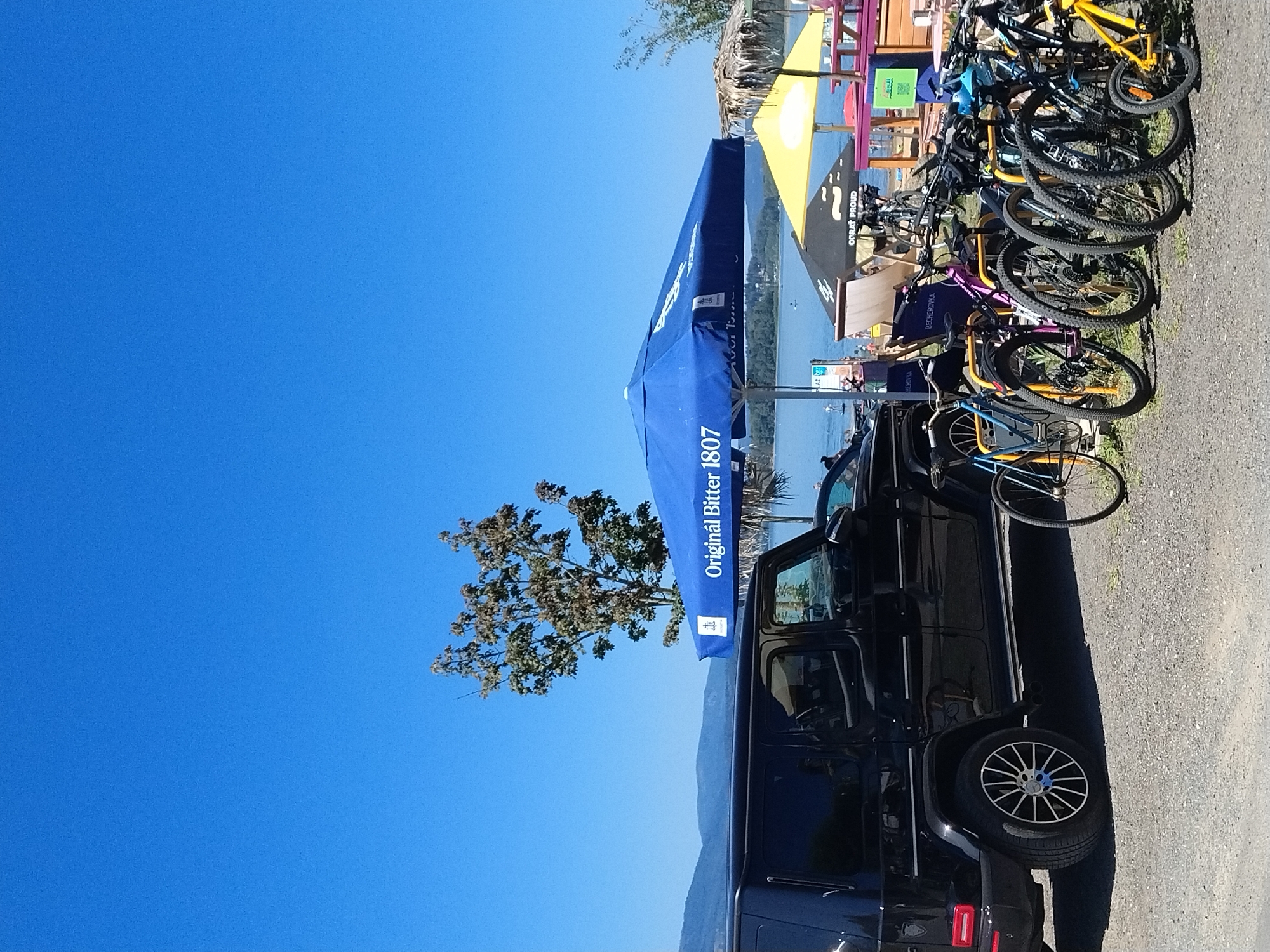 The image depicts an outdoor scene, likely a lakeside area, under a clear blue sky. In the foreground, a black Mercedes-Benz G-Class vehicle is parked on a gravel surface. To the right of the vehicle, a row of parked bicycles of various colors is visible. Several umbrellas, one large blue one with the text "Originál Bitter 1807", and a yellow one, are set up, suggesting a recreational or dining area. In the background, a body of water is present, with some people visible on or near the water, possibly engaged in water sports. Hills or mountains can be seen in the distance, forming a natural backdrop. The overall impression is one of a sunny day at a popular outdoor location. The location is stated to be Radslav, Czechia, which suggests it is likely a lake or dam area in or near that municipality, possibly associated with a resort or recreational facility.