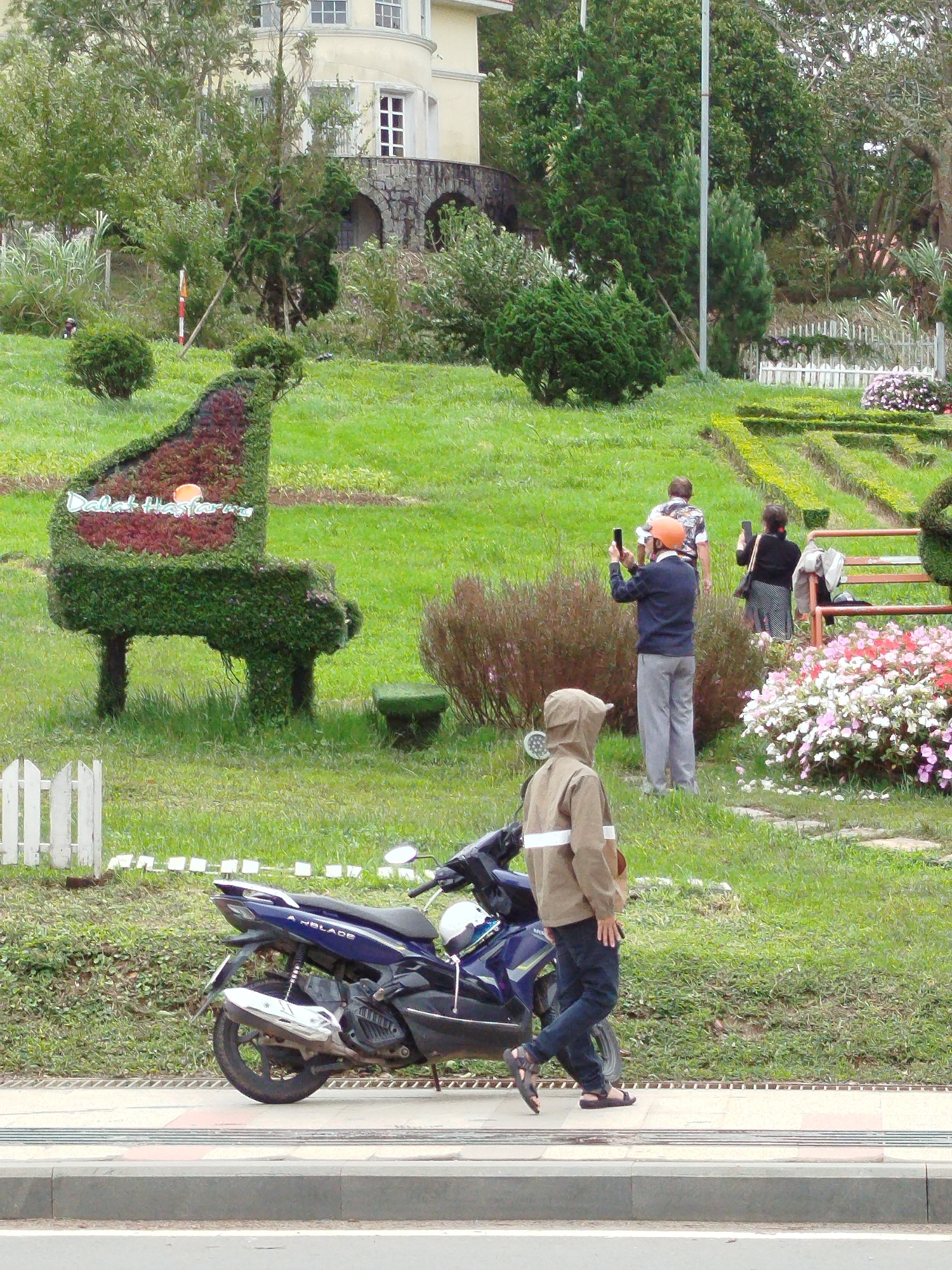 The image depicts a park-like setting in Da Lat, Vietnam. In the foreground, a person wearing a brown hooded jacket and dark pants walks towards the left, alongside a parked blue motorcycle. The motorcycle has "A BLADE" written on its side. In the middle ground, there is a topiary shaped like a grand piano adorned with red flowers and green foliage, with "Dalat Hasfarm" written on it. Behind the piano topiary, a man in a blue shirt and grey pants takes a photo with his phone, while a woman in a black top and patterned skirt also holds a phone. Further back, another person is visible walking away. The background features lush green grass, trees, and a building with arched stonework. White picket fences and flowerbeds are also present in the scene. The overall atmosphere suggests a tourist destination or a well-maintained public garden.