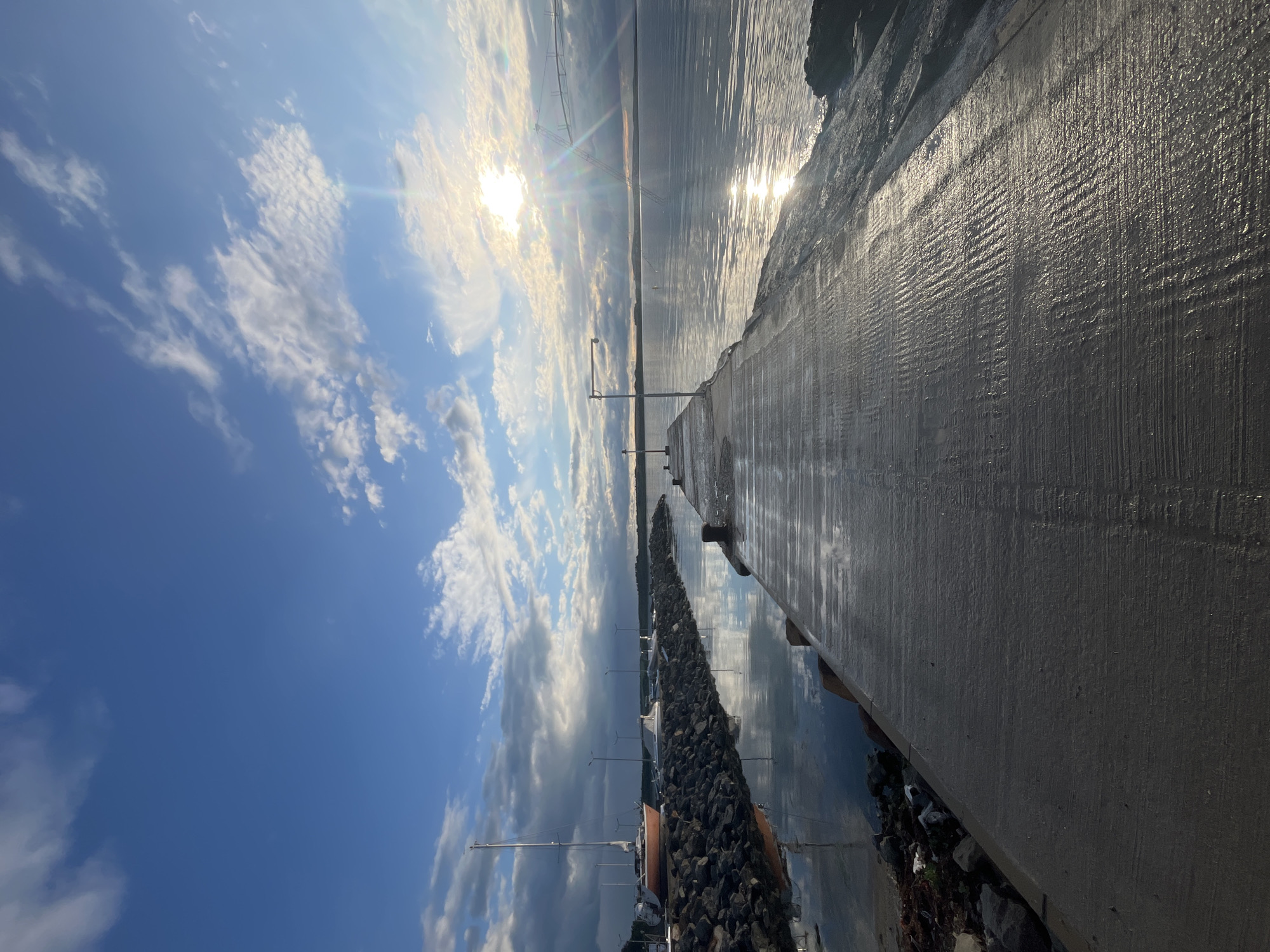 The image shows a concrete pier extending into the Black Sea in Burgas, Bulgaria. The sky is blue with white clouds and the sun is reflecting brightly off the water. A line of rocks runs along the edge of the pier, bordering a marina with several boats. The pier appears wet, suggesting it has recently rained or been washed by waves. There are no people visible.