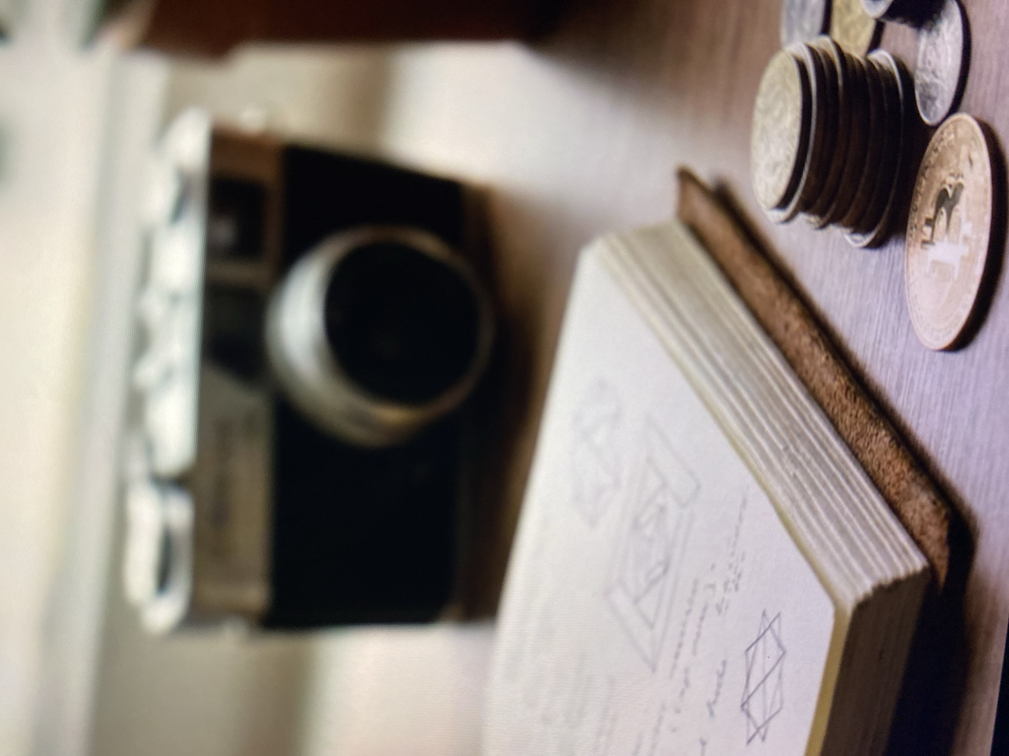 The image depicts a still life arrangement on a wooden surface. In the background, a vintage camera, likely a rangefinder, is out of focus. In the foreground, a closed notebook with a leather cover is placed on the surface. The notebook's pages are visible, and they appear to have sketches and handwritten notes. To the right of the notebook, a stack of coins is piled, with some individual coins scattered around them. One of the coins in the foreground prominently displays the Bitcoin logo. The overall lighting suggests an indoor setting, possibly near a window. There are no people or any indication of a specific location in Kuta Selatan, Indonesia.