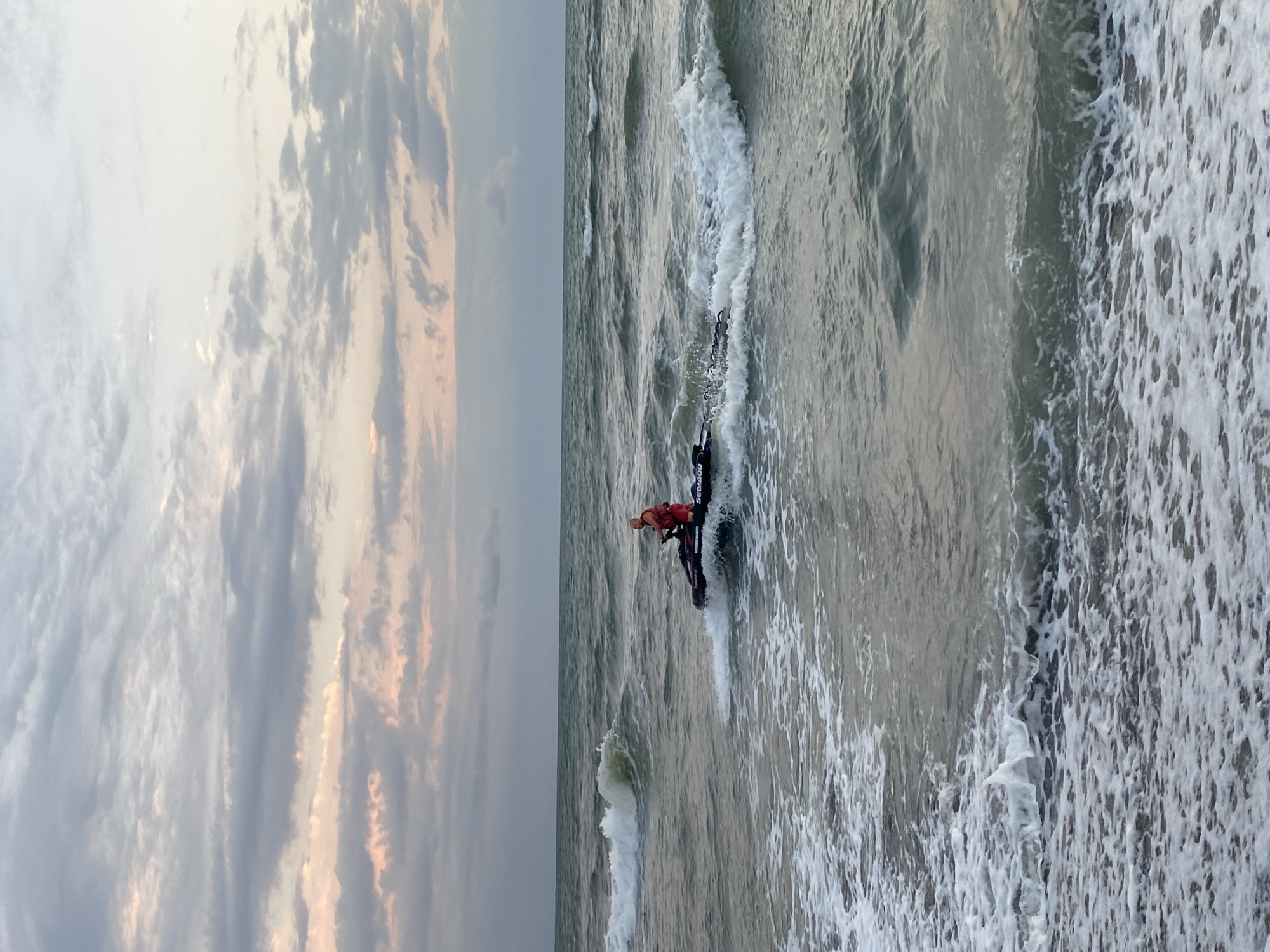 A man wearing a red life vest and red shorts is riding a black and purple jet ski on a choppy sea. The jet ski is creating a white spray as it moves through the water. The sky above is filled with dramatic, multicolored clouds, with hues of pink, gray, and white, suggesting either sunrise or sunset. The sea is a muted green-gray color, with white foam from the waves breaking near the shore. The image appears to have been taken from a beach. The location is Ureki, Georgia.
