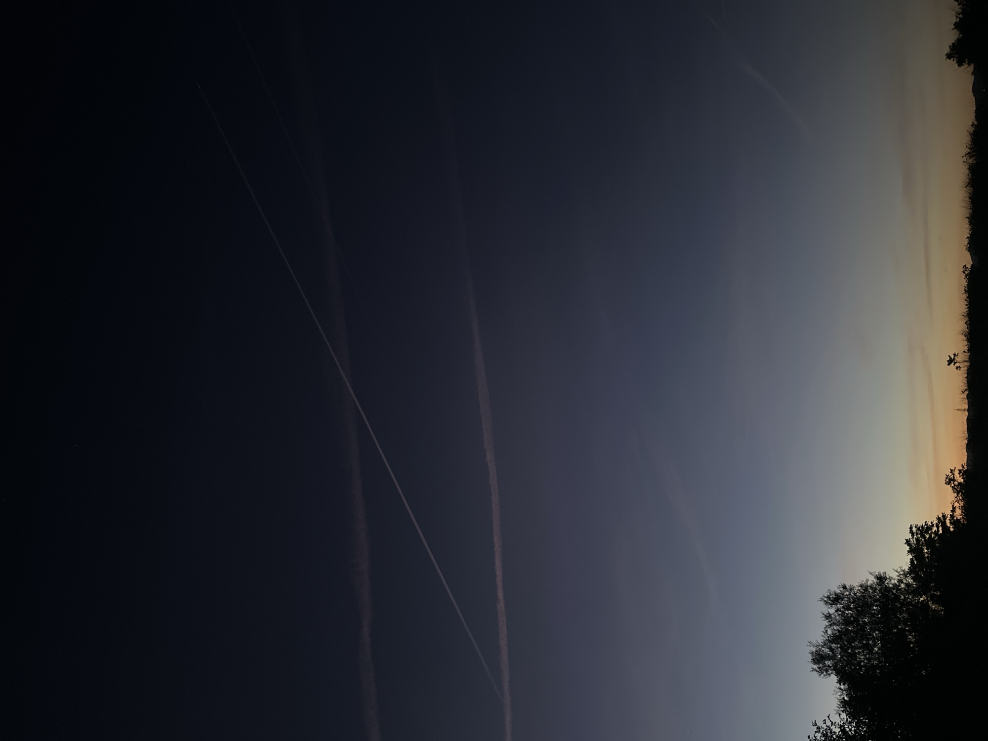 The image depicts a dark sky at dusk or dawn, with contrails from aircraft visible. The darkness fades from left to right into a lighter band on the right where the horizon is visible, marked by silhouettes of trees. The angle of the photograph implies it was taken from a position in a relatively open area, perhaps a park or field. There is not enough data to determine the exact location, and there is no information to suggest this photo was taken in Sofia, Bulgaria. Further detail would require additional contextual clues or visible landmarks. There are no people visible.