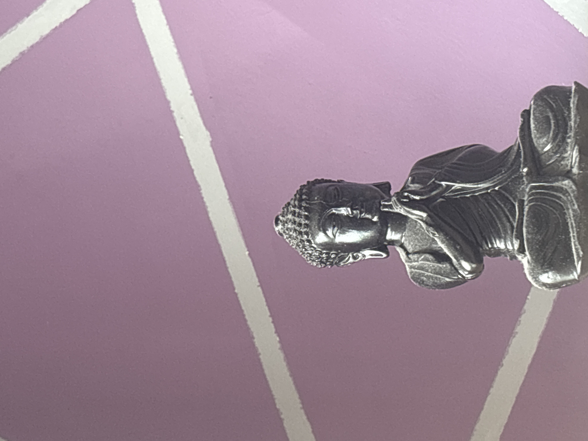 A close-up shot depicts a metal Buddha figurine oriented horizontally, with its head positioned toward the left side of the image. The figurine rests against a light purple background marked with broad white stripes that converge in a geometric pattern. The details of the Buddha figurine are sharp, revealing the texture of its hair and the folds of its robes. The setting does not appear to be in Selyanin, Bulgaria.
