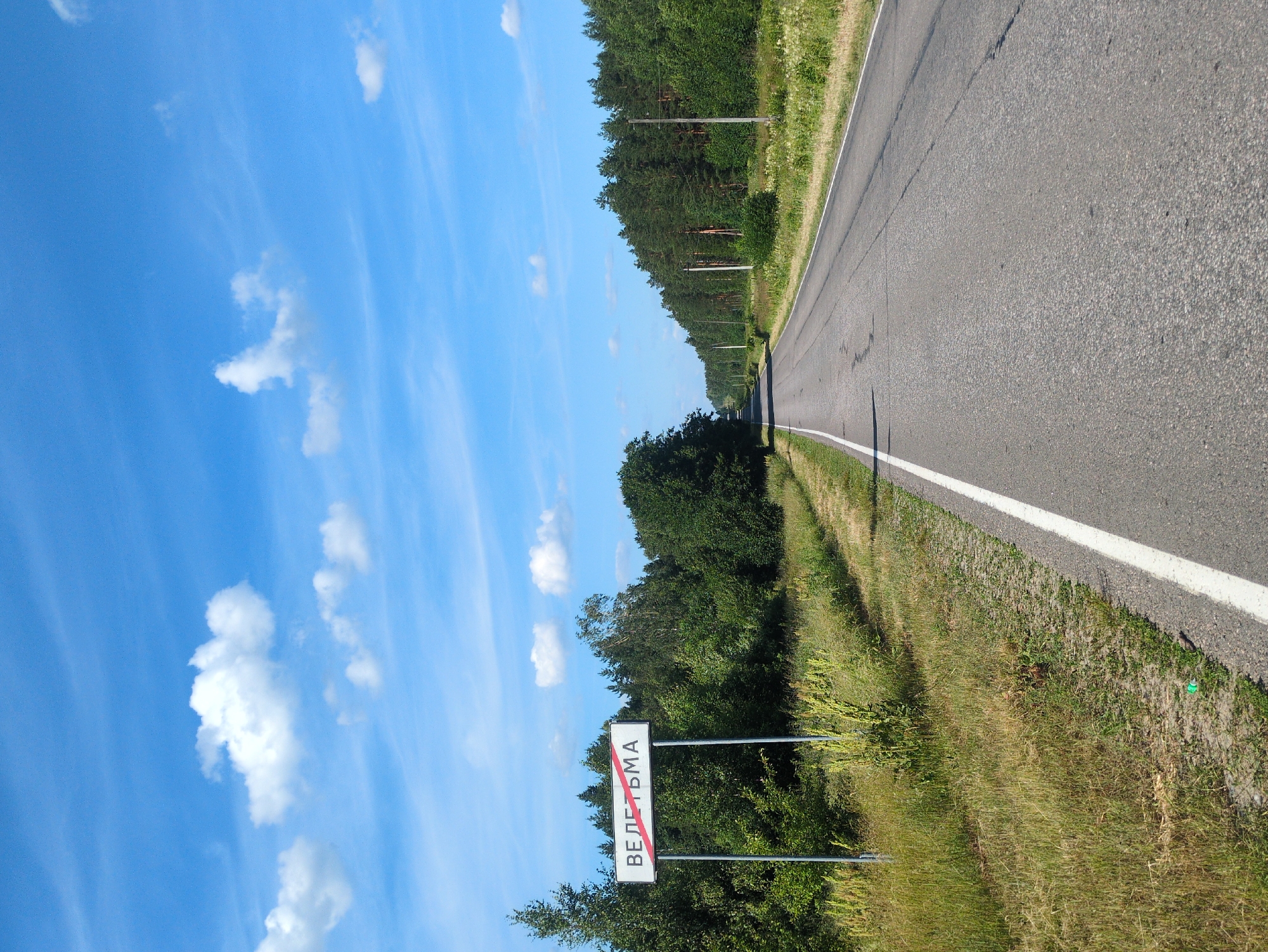 A road sign reading "ВЕЛЕТЬМА" with a red diagonal line through it, indicating the city limits of Velet'ma, Russia, is visible on the left side of a paved road. The sign is white with black lettering and is mounted on two metal poles. The road stretches into the distance, flanked by lush green trees and vegetation. The sky above is a clear blue with scattered white clouds. The scene is a rural landscape on a sunny day.