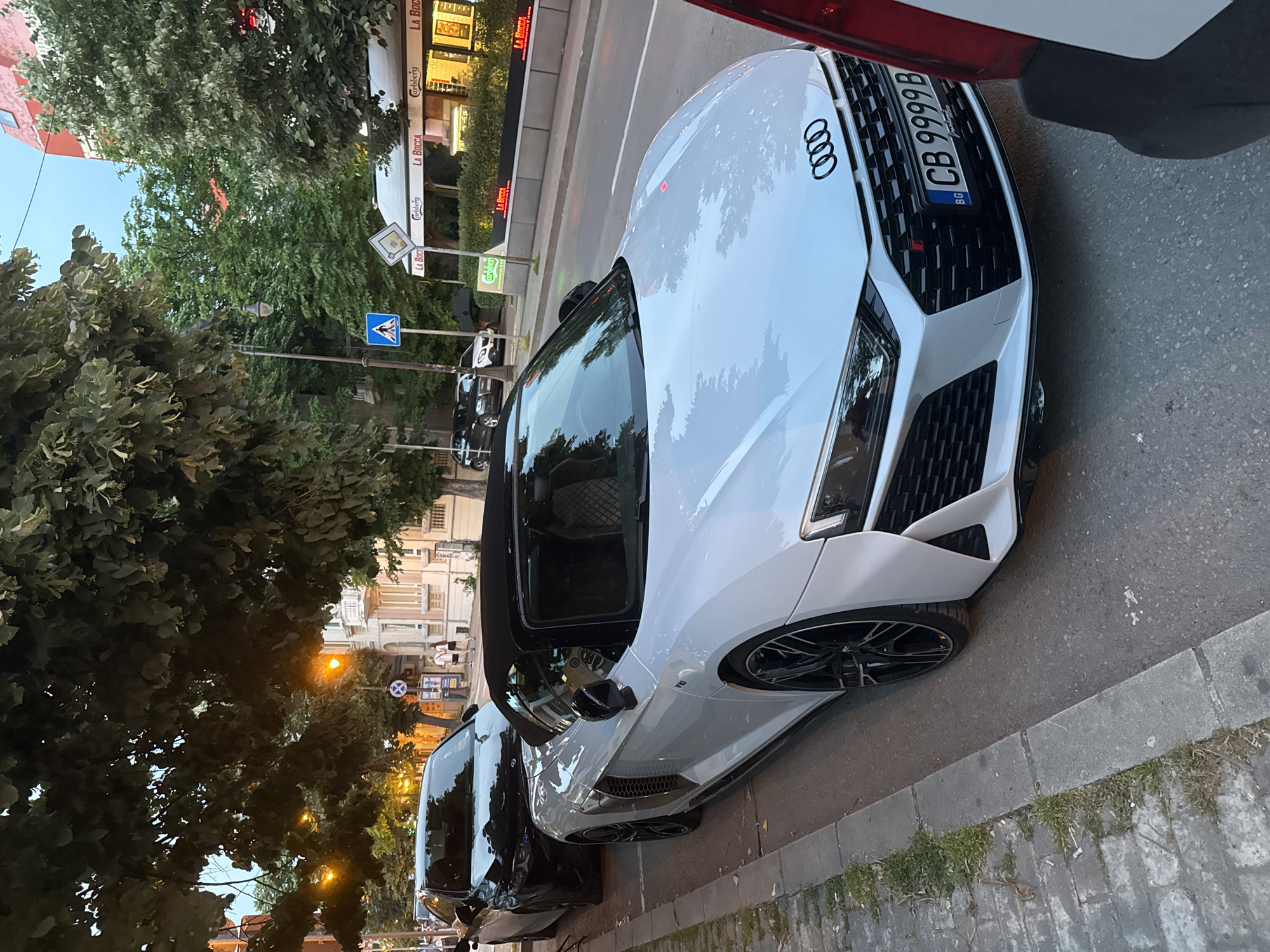 A white Audi R8 convertible is parked on a street next to a sidewalk. The car has its black convertible top down and its front license plate is visible, reading "CB 9999 B". A black car is parked behind the Audi. Trees with green leaves overhang the street and sidewalk. Buildings with windows and balconies are visible in the background, along with streetlights and signs for businesses, including "La Bocca" and "Carlsberg". The sky is a light blue, suggesting it is daytime.
