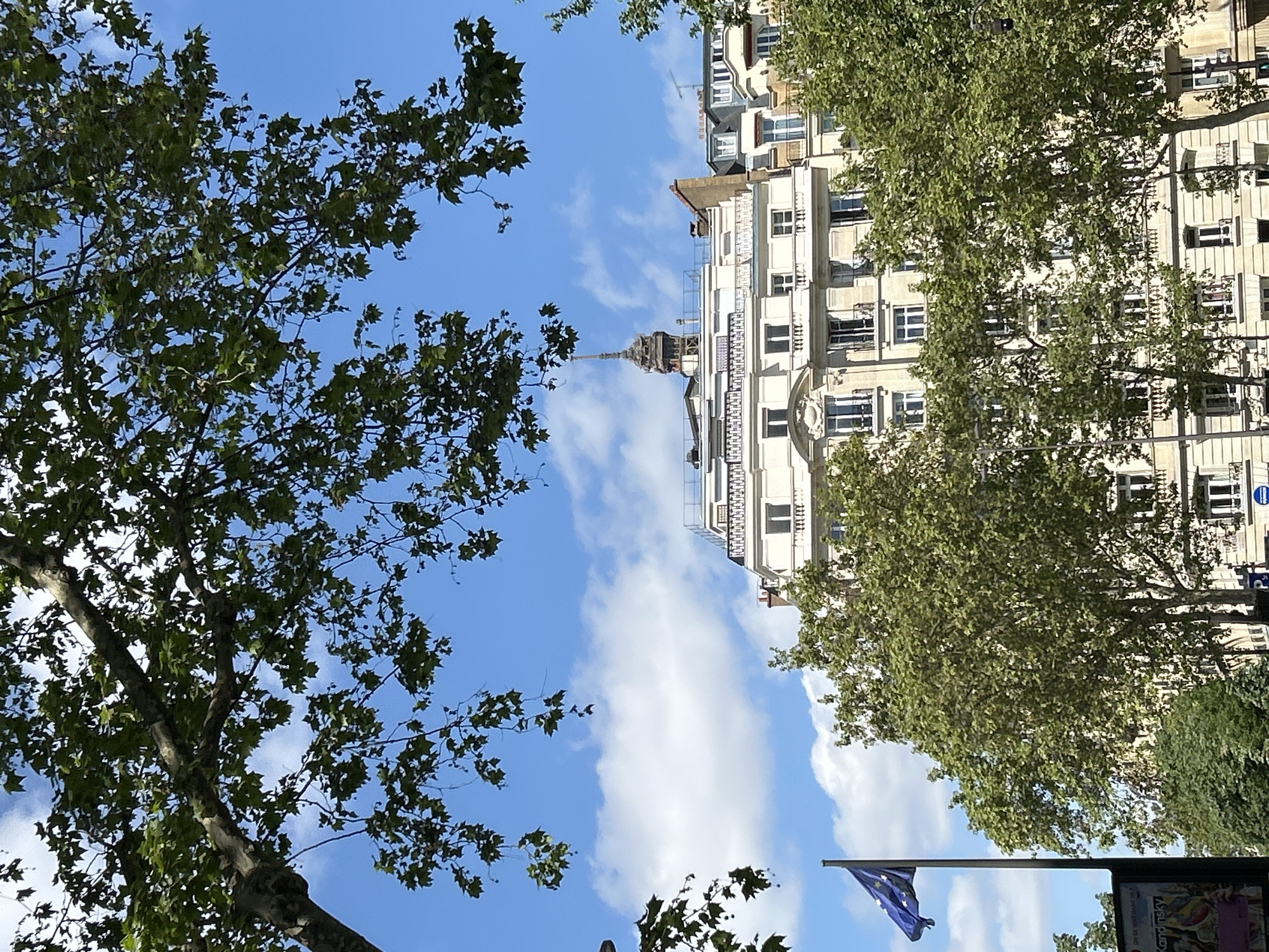 The image captures an upward-looking view of a city scene under a bright blue sky dotted with fluffy white clouds. Lush green trees, likely plane trees with their distinctive foliage, frame the left and right sides of the composition, their branches extending towards the center.
In the middle-ground, a grand, multi-story building of light-colored stone or stucco dominates the right side. It features numerous rectangular windows, many with balconies adorned with wrought-iron railings. The architectural style suggests a classic European design, possibly Haussmannian.
Centered in the background, peeking above the rooftops of the buildings, is a tall, slender spire or cupola. Its intricate dark structure contrasts with the light sky, giving a sense of depth and hinting at a significant landmark in the distance, possibly the top of the Eiffel Tower or another prominent city spire.
In the bottom right corner, a blue flag with yellow stars, identifiable as the European Union flag, flies from a pole. Below it, part of a black street furniture object, possibly an advertising column or information stand, is visible. Text on this object clearly shows the name "Kandinsky."
The scene is bathed in natural daylight, indicating a sunny or partly cloudy day. There are no people or active events depicted, creating a serene and architectural cityscape view. The combination of the distinctive architecture, the European Union flag, and the potential spire suggests a major European capital, likely Paris.