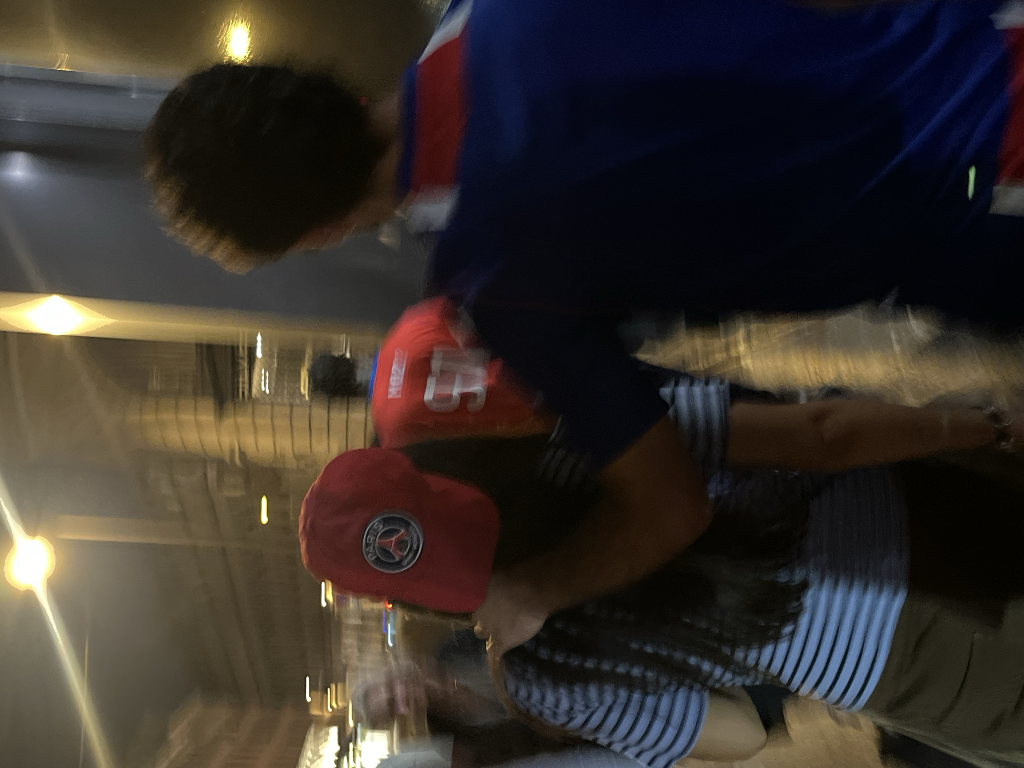 A blurry, vertically oriented image depicts two individuals in an outdoor setting, potentially in Paris, France. The individual at the top wears a dark blue shirt with red and white accents. Below them, another person is wearing a red baseball cap featuring a visible Paris Saint-Germain logo and a striped shirt with a possible white and blue pattern. The surrounding environment is poorly lit, appearing to be a street lined with buildings. It's difficult to ascertain specific actions or interactions due to the image's lack of clarity.