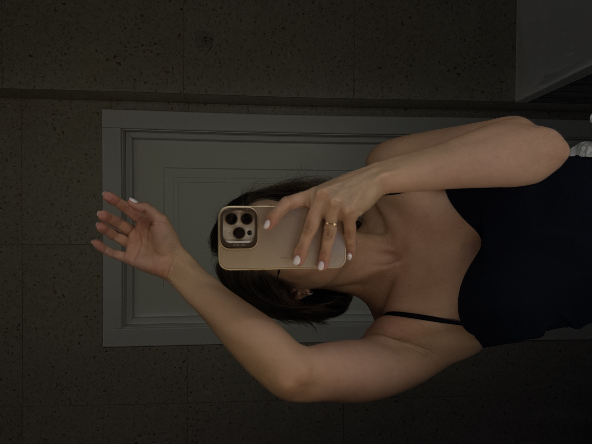 A woman is taking a selfie in a bathroom. She is holding a gold iPhone in her right hand, obscuring her face. She is wearing a black tank top. Her fingernails are painted white. She is wearing a gold ring on her left ring finger. The background consists of tiled walls and a door. It's not possible to tell the location is Pervomaysky, Russia, based on the image.