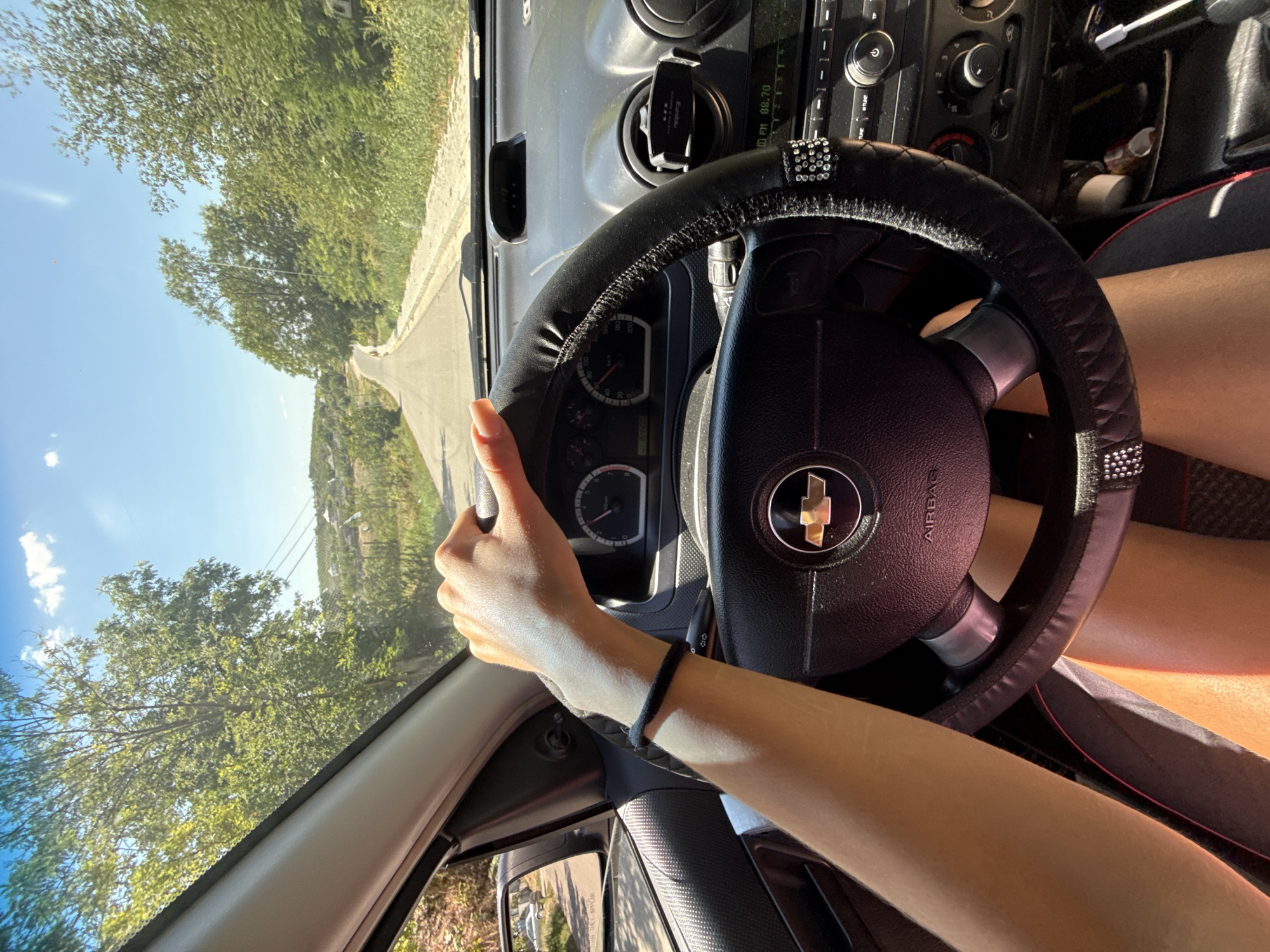 A first-person perspective shot captures the scene from inside a car, focusing on the driver's hands on the steering wheel. The vehicle is on a paved road that winds uphill through a rural, verdant landscape. Trees line both sides of the road, with a small cluster of houses visible in the distance on a hilltop. The sky is clear blue with a few wispy clouds. The car's dashboard, including the steering wheel with a Chevrolet emblem, the instrument cluster, and the radio, is clearly visible. A person's arm, with a black wristband, rests on the steering wheel. The overall impression is of a sunny day drive through the countryside, possibly in or around Basarabeasca, Moldova.