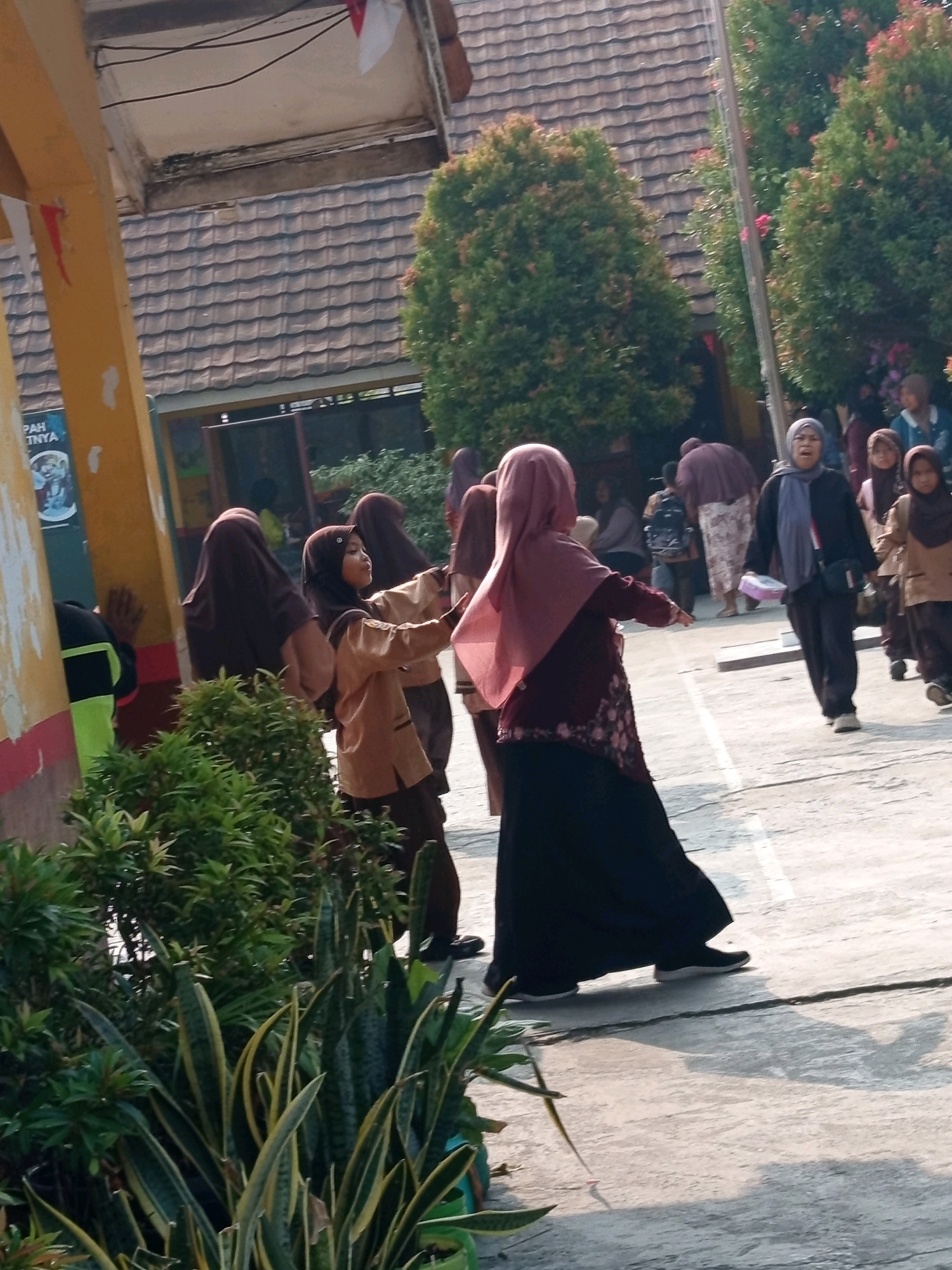 The image depicts an outdoor scene in Karawang, Indonesia, likely at a school. Several young girls wearing hijabs and school uniforms are visible, some engaged in what appears to be a group activity or game. One girl in the foreground, dressed in a tan uniform, is reaching out, while another in a maroon hijab and skirt is facing away from the camera. Behind them, other individuals, including adults and more children, are present. The environment includes a building with a tiled roof, greenery such as trees and potted plants, and a paved outdoor area. The lighting suggests it is daytime.