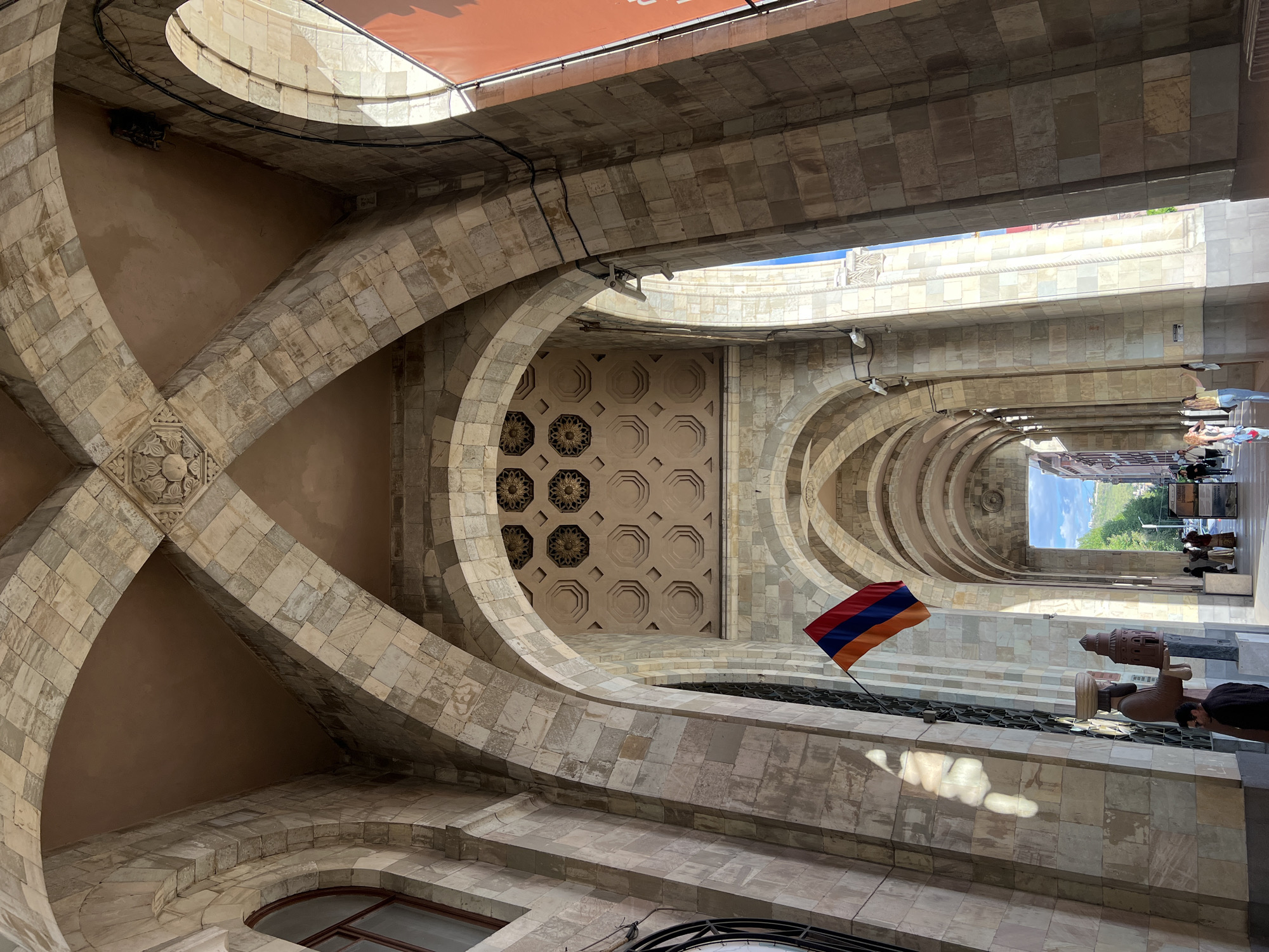 The image shows an interior view of a building in Yerevan, Armenia. The architecture features arched ceilings and stone walls. An Armenian flag is visible. In the background, there is a view of the outdoors, showing a street scene with people and trees.