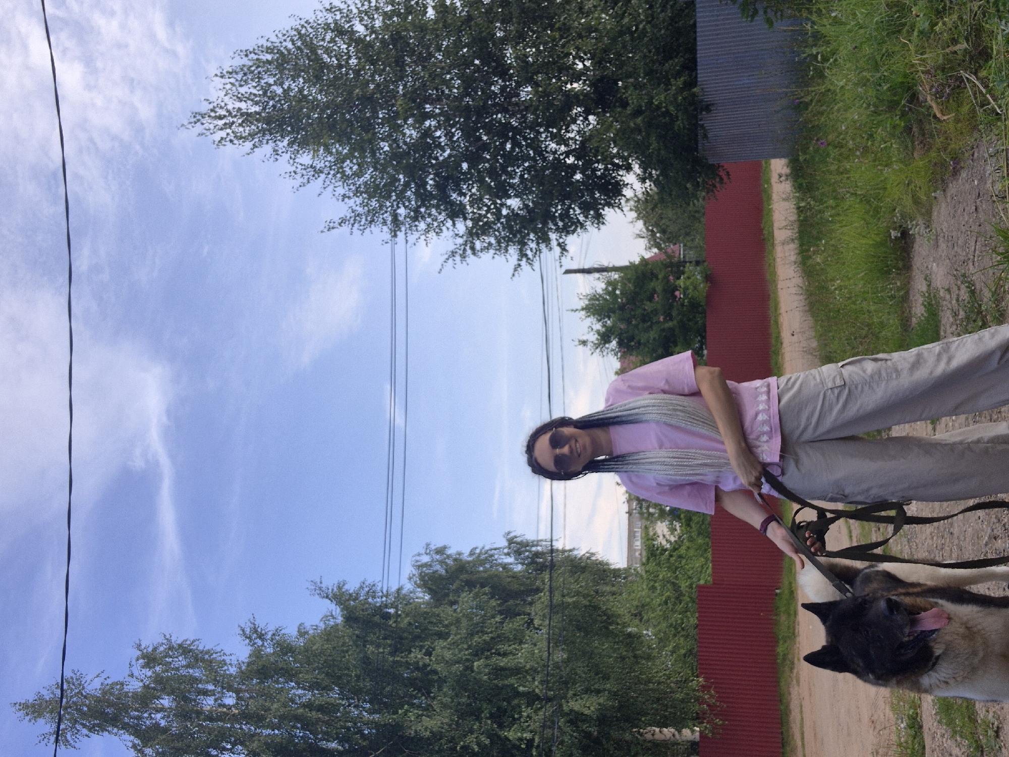 A person stands outdoors holding a dog on a leash. The person is wearing a pink t-shirt, beige cargo pants, and sunglasses. They have long, braided hair. The dog is a large breed, possibly an Akita, with black and tan fur. The background features trees, a red fence, a corrugated metal fence, and a dirt path. The sky is blue with some clouds. The scene appears to be in a rural or suburban area.