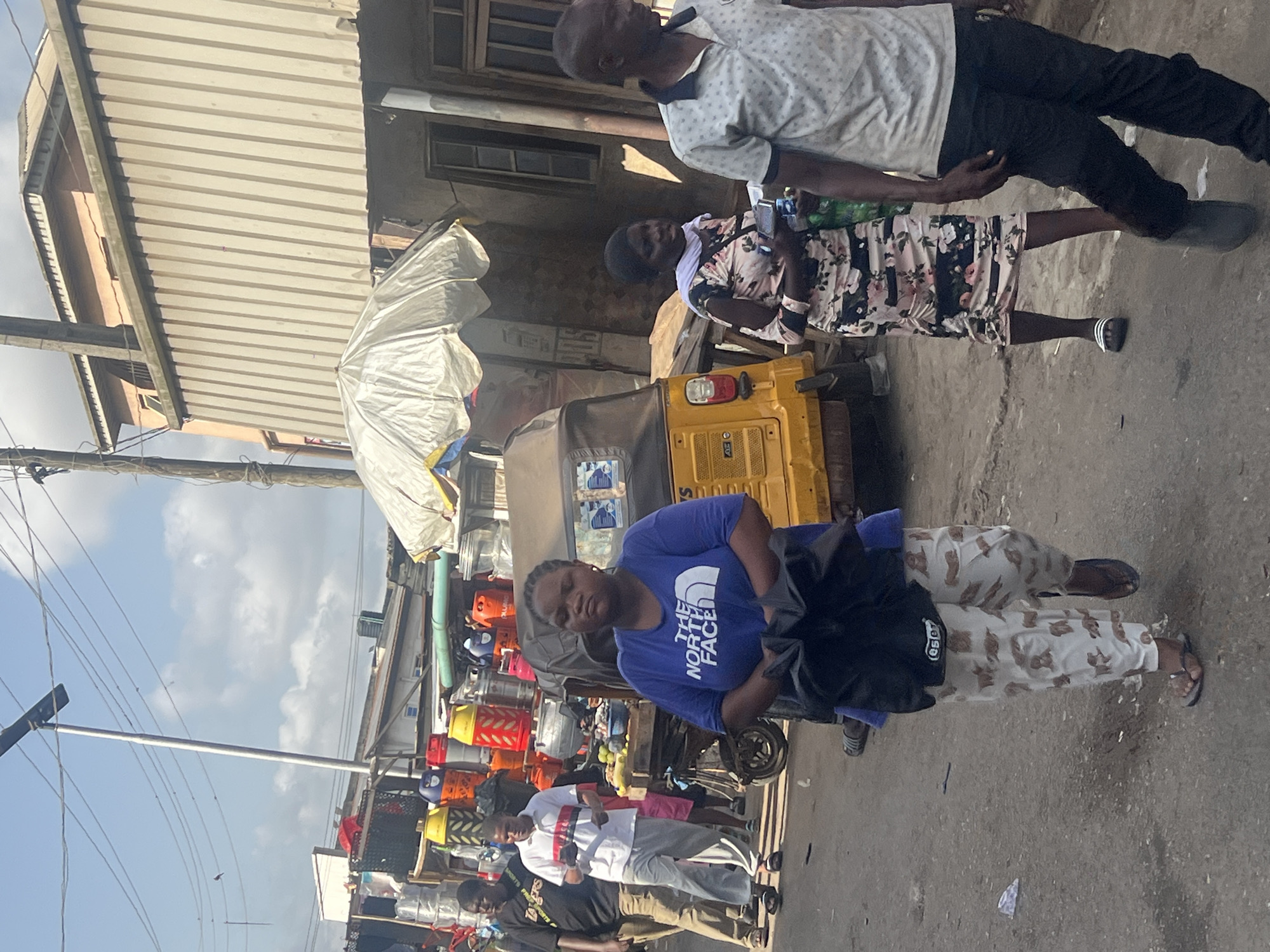 The image shows a street scene in Egan, Nigeria. In the foreground, a woman wearing a blue "The North Face" t-shirt and patterned pants stands on the right. To the left of her are stacks of colorful gas cylinders. Behind her is a yellow auto rickshaw. To the right, an older man and woman stand in the street. The man wears a white spotted shirt and black pants, and the woman is wearing a floral dress. In the background, a corrugated metal roof is seen on the left. There is a streetlight on the left side of the image. People walk along the street on the left.