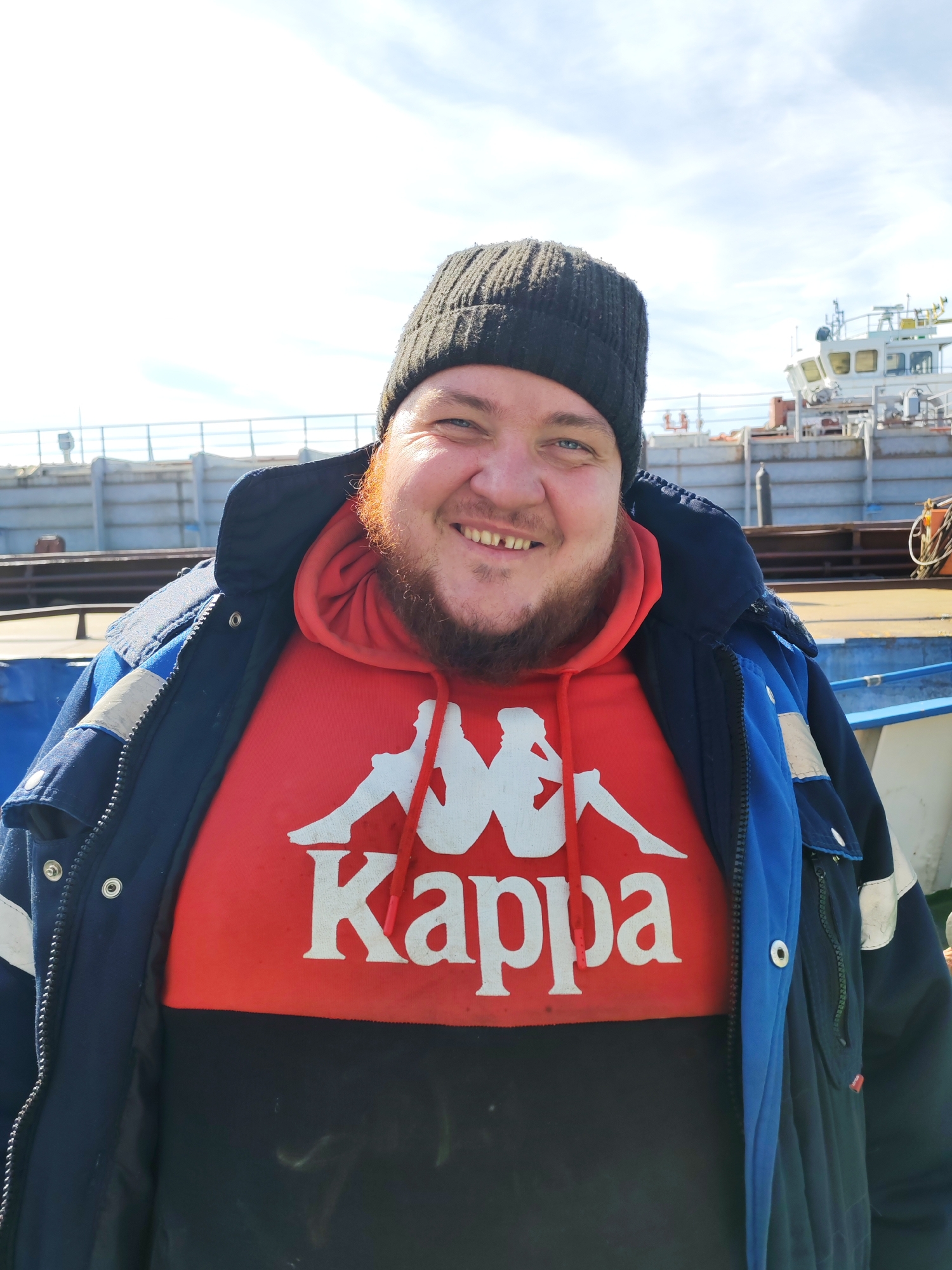 A smiling man is featured in a close-up, chest-up portrait, standing outdoors on what appears to be the deck of a large vessel. He wears a dark grey or black knitted beanie, a red and black Kappa brand hoodie, and a heavy-duty blue and black work jacket. His reddish-brown beard is full, and he smiles broadly, showing a gap between his front teeth. His eyes are blue.
The red section of his hoodie prominently displays the white "Kappa" text along with the brand's iconic logo of two figures leaning against each other. The blue and black jacket is open, revealing the hoodie, and features functional details like a zipper, snaps, and what appears to be a reflective strip on the left arm.
The background shows the superstructure of a ship or large boat, with white and grey sections, and various equipment like railings and ropes. The sky above is bright and partly cloudy, suggesting it is daytime. The man appears cheerful and comfortable in his attire, set against a working maritime environment.