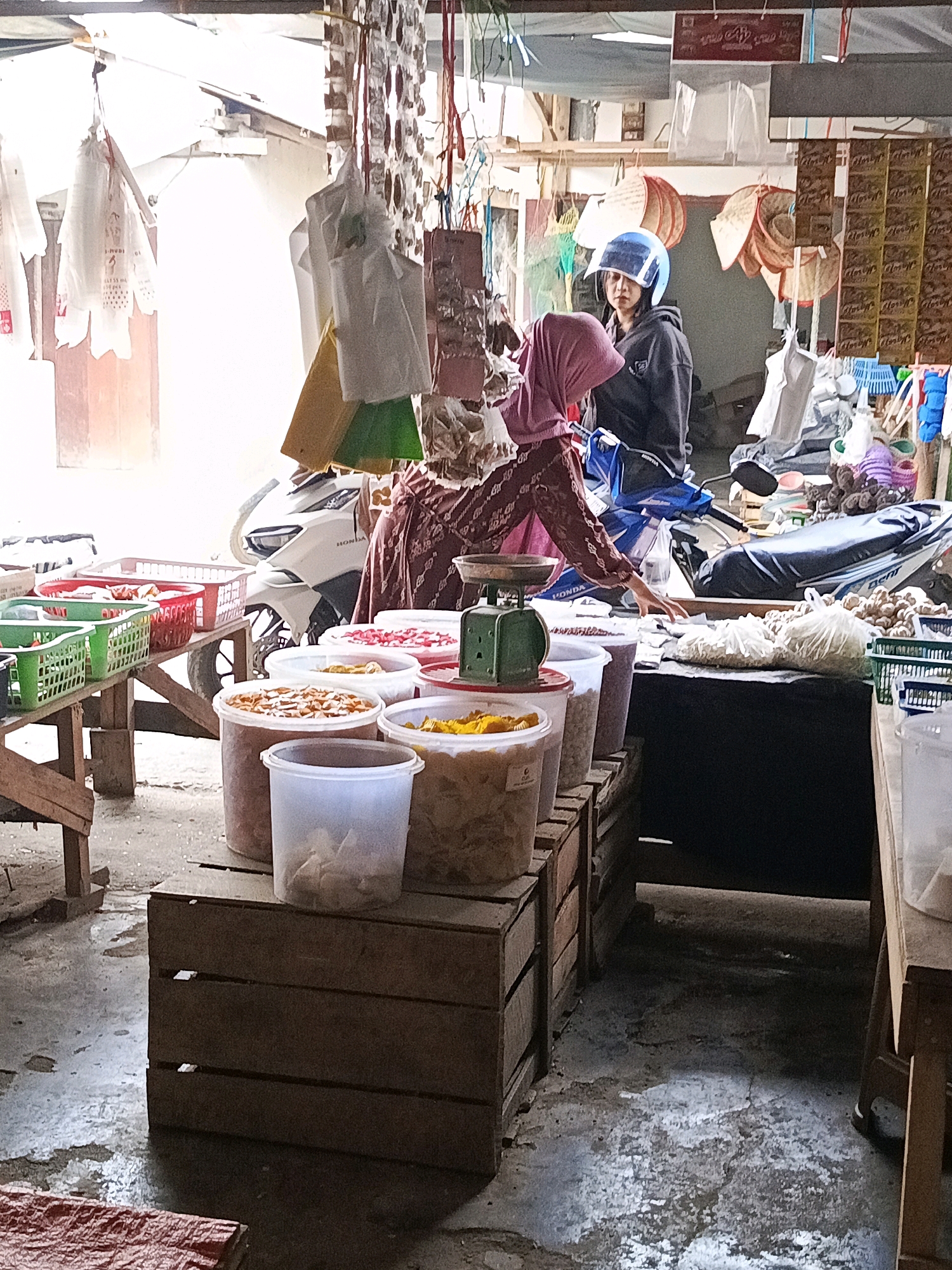 The image depicts an outdoor market stall, likely in Megang Sakti I, Indonesia, on a bright day. Two individuals are present. The person in the foreground, wearing a pink headscarf and a patterned brown top, appears to be a vendor. They are reaching towards a digital scale on a table laden with various food items in plastic containers, including what appear to be snacks or dried goods in shades of orange and yellow. Behind them, a younger individual wearing a blue helmet and a dark jacket is seated on a motorcycle, looking towards the front of the stall.
The stall itself is a chaotic yet organized collection of goods. To the left, wooden shelves hold red and green plastic baskets filled with packaged items. A white motorcycle is parked behind the individuals, partially obstructing the view. Various items are suspended from above, including plastic bags, what resemble woven fans, and other market wares. The ground appears to be concrete, with some scattered debris and puddles reflecting the overhead light. The overall impression is of a busy, functional marketplace.