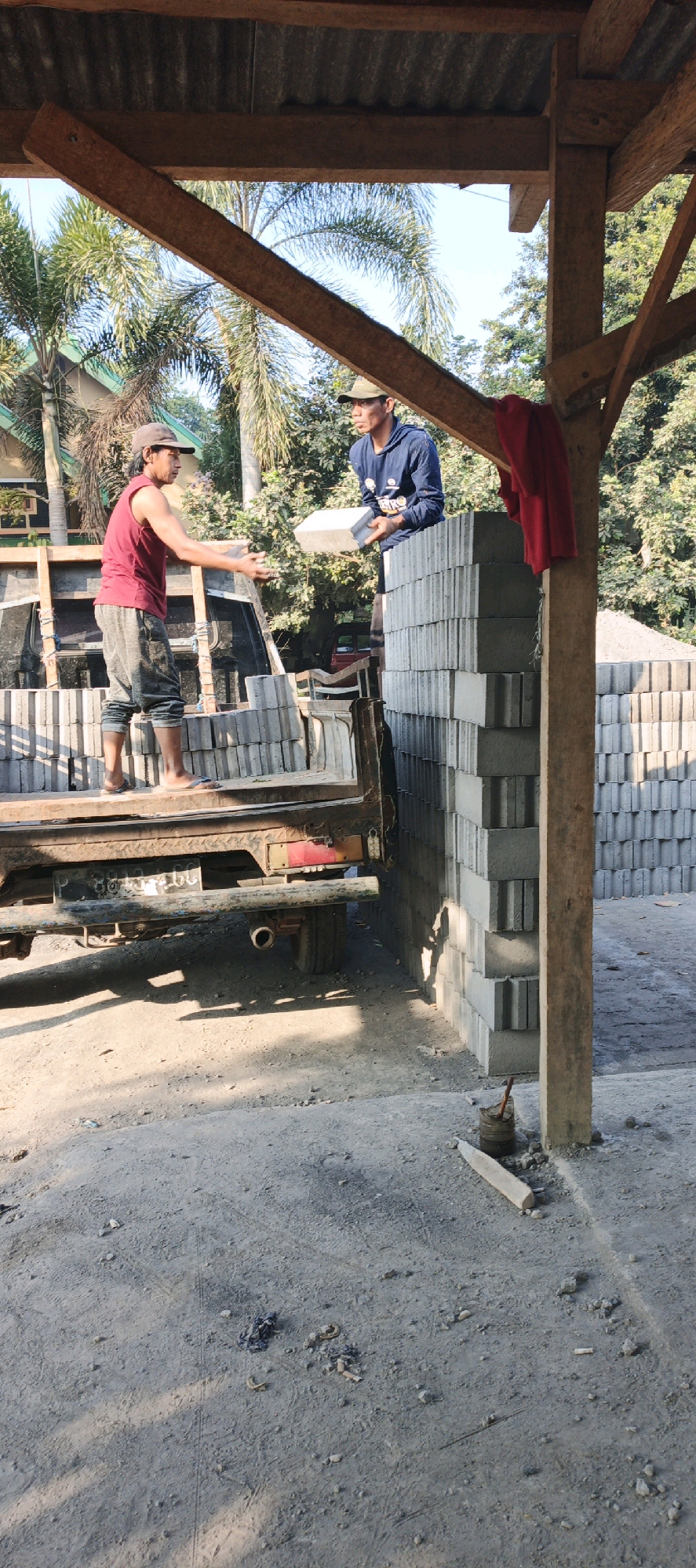 Two men are working under a wooden shelter, possibly a construction site or storage area, in Kasiyan, Indonesia. One man, wearing a red sleeveless shirt and grey shorts, stands on the bed of a truck, extending his hands towards the other man. He appears to be receiving or handing off a brick. The second man, dressed in a blue hooded sweatshirt and a baseball cap, stands on a tall stack of gray concrete bricks, holding a brick in his hands. The truck bed is also filled with bricks. In the background, lush green trees and palm trees are visible, suggesting a tropical environment. A red cloth is draped over a wooden beam of the shelter. The ground is dirt and gravel, with some debris scattered around. The scene depicts a task involving the handling and stacking of construction materials.