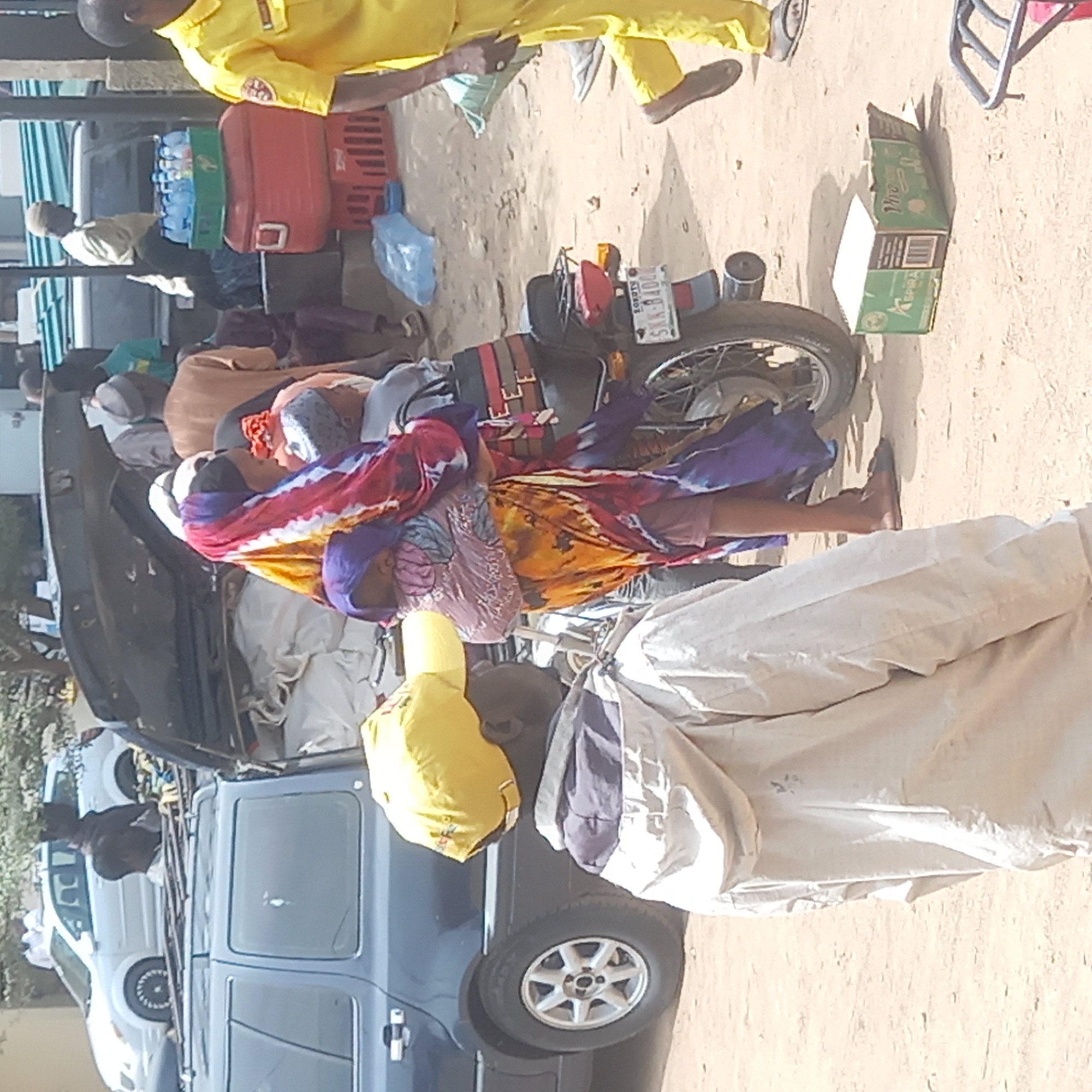 The image depicts a scene in what appears to be a busy outdoor area in Dadi Riba, Nigeria. A dark-colored SUV is packed with items on its roof, including bags covered with tarps, suggesting people are travelling or transporting goods. A woman dressed in colorful robes stands next to a motorbike loaded with luggage, secured with straps. The motorbike has a Nigerian license plate that reads "SNA-840LE." In the background, other vehicles are visible, packed with people and goods. A man wearing a yellow uniform stands nearby, possibly a traffic warden or a member of a security service. The setting appears to be a sandy area with buildings partially visible in the background. There is a carton on the ground next to the motorbike.