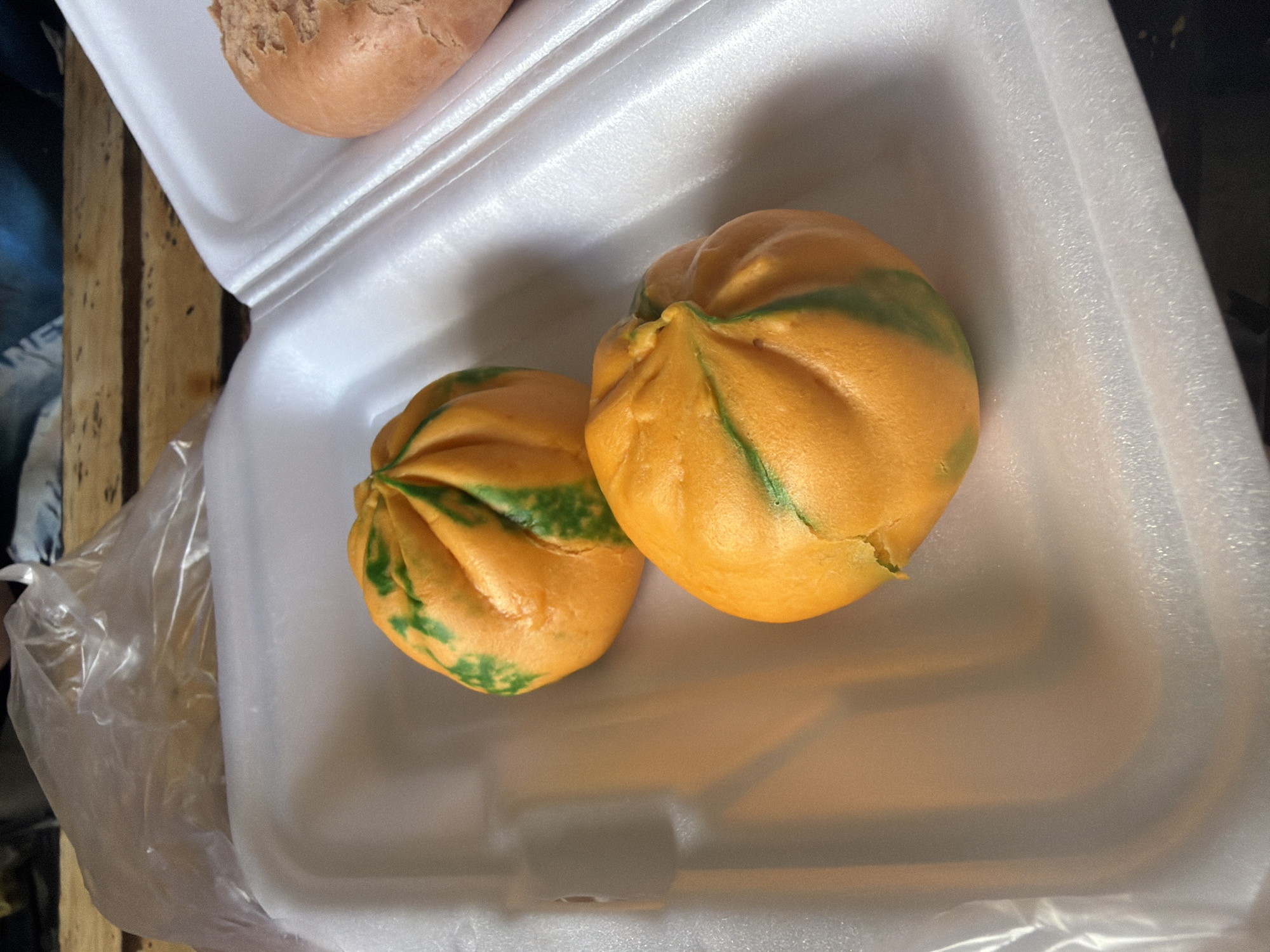 This image captures a close-up view of food items packaged in a white styrofoam container, likely for takeout or sale in Măng Đen, Vietnam.
The primary subjects are two brightly colored buns, appearing to be steamed buns (like *bao* or *bánh bao*). They are predominantly orange with striking, irregular green streaks and patches, creating a marbled or patterned effect. Both buns are round and plump, with visible folds in their dough, particularly at the top where they seem to have been sealed.
Partially visible in the upper left corner of the container is a third bun or piece of bread, which is a uniform light brown color and has a rough, textured surface, different in appearance from the orange and green buns.
The buns are neatly placed within the white styrofoam container, which shows some wear and minor indentations. To the left of the container, a clear, crinkled plastic bag is visible, partially obscured by the container's edge. Below and to the far left, a wooden surface, possibly a table or crate, with a rustic grain and dark specks, serves as the background.
The scene depicts food ready for consumption or purchase. The style of buns and packaging are consistent with street food or market offerings common in Vietnam. There are no people visible, no discernible activity beyond the presentation of the food, and no visible text. The lighting is soft and even, suggesting either an indoor setting or a shaded outdoor area.
