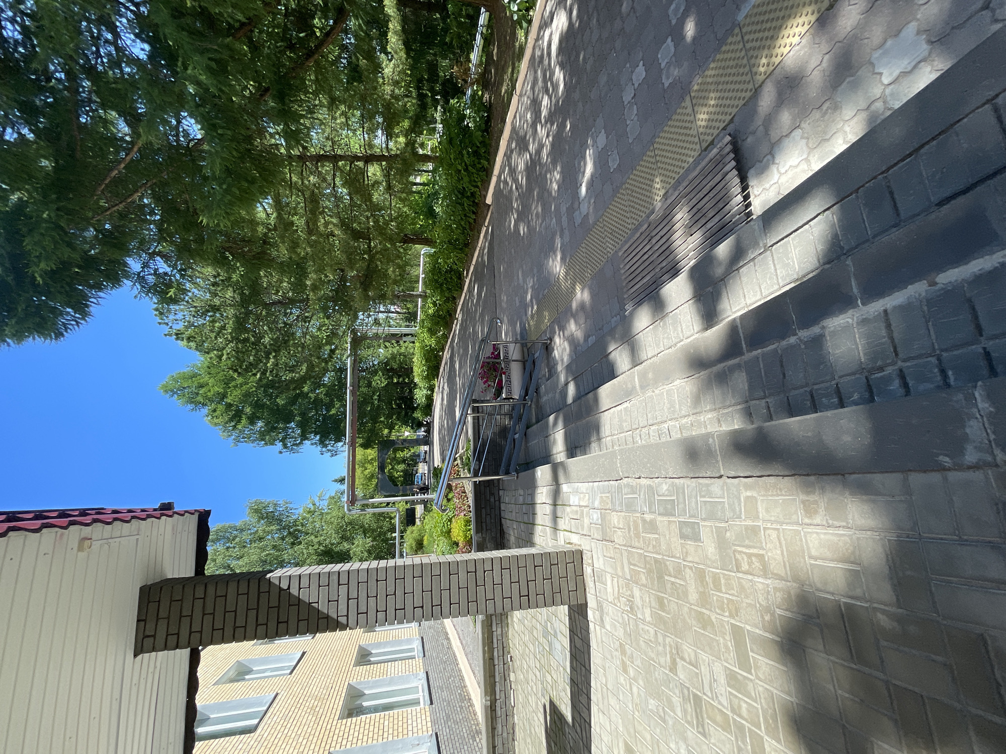 The image shows a paved walkway leading up to a building with a light yellow brick facade and a white soffit with a red decorative trim. A dark grey brick pillar stands to the left of the walkway, casting a shadow on the light grey and beige patterned paving stones. The walkway includes a metal handrail and a ramp with stairs. Further along the walkway, there is a wooden slatted bench and tactile paving for visually impaired pedestrians. The scene is bordered by lush green trees and bushes, and the sky is a clear blue. In the background, there is an arched structure and a glimpse of a car. The lighting suggests it is daytime, with strong sunlight creating distinct shadows. The location is identified as Krasnye Baki, Russia.