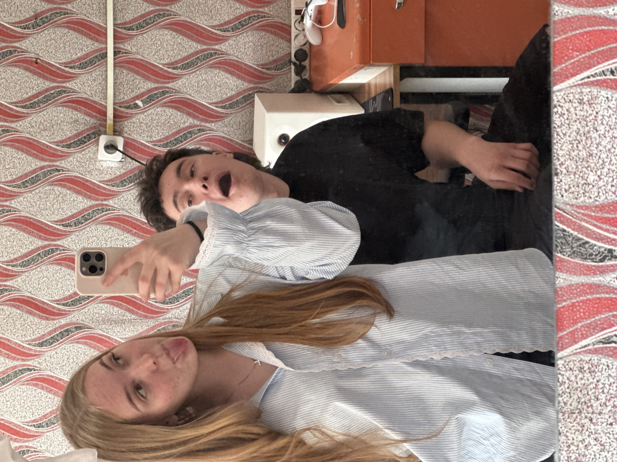 A selfie is taken by a fair-skinned young woman with long blonde hair and blue eyes wearing a light blue striped shirt with lace trim. She is sticking her tongue out. Behind her is a dark-haired male wearing a black shirt with his mouth open. The phone held by the woman is a beige color. The location appears to be inside a building in Perm, Russia, as evidenced by the red-and-gray patterned wallpaper and a European-style electrical outlet. Behind the man is what appears to be an orange cabinet, a white speaker, and a reflection in a mirror.