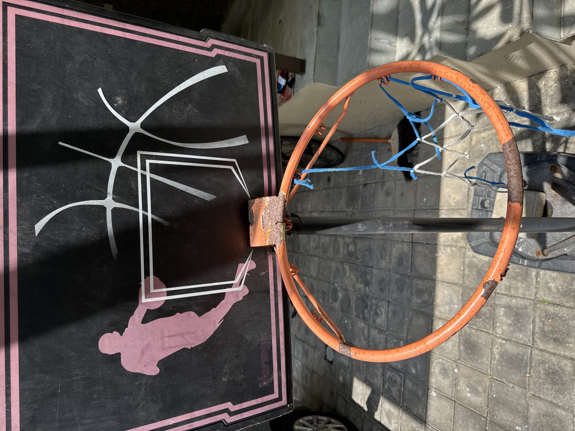 The image depicts a worn basketball hoop outdoors. The backboard is black with a pink border, featuring a graphic of a basketball player and white lines resembling the court markings. The hoop itself is orange and shows signs of rust and wear, with a makeshift net made of blue and white ropes. The scene appears to be in an outdoor setting with tiled pavement. Although the precise location within Jimbaran, Indonesia is not discernible from this single image, it is clearly an outdoor recreational area or private residence in this city.