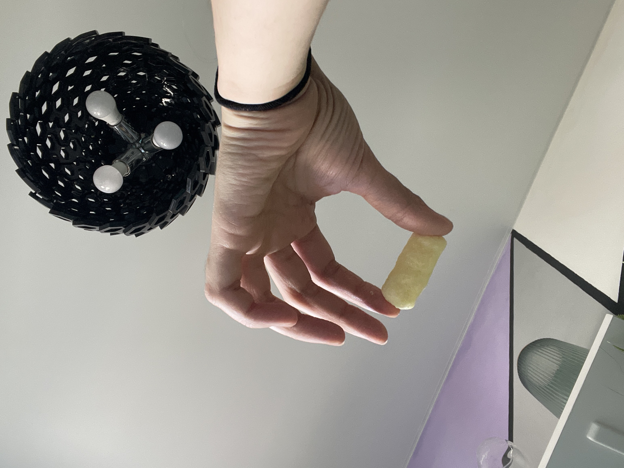 A low-angle shot captures a person's hand holding a yellow, cylindrical object between their thumb and forefinger. The hand has a black wristband. Above and to the left of the hand is a black, modern-style light fixture with three visible light bulbs. The background consists of a white ceiling, with a portion of a wall with a lilac section visible on the right. A modern painting or decorative object with geometric shapes and a green element is partially visible in the lower right corner. The location is an interior space in the city of Unknown, Unknown.