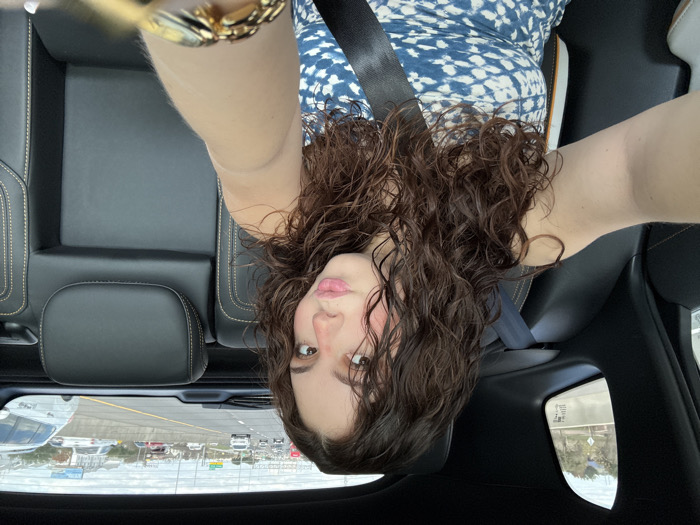 A woman with curly brown hair and a blue and white patterned dress is sitting in a car. She is wearing a seatbelt. Her head is tilted back, and she is looking at the camera. The view out the window shows a highway with cars. The car's interior is black. A gold watch is visible on her wrist.